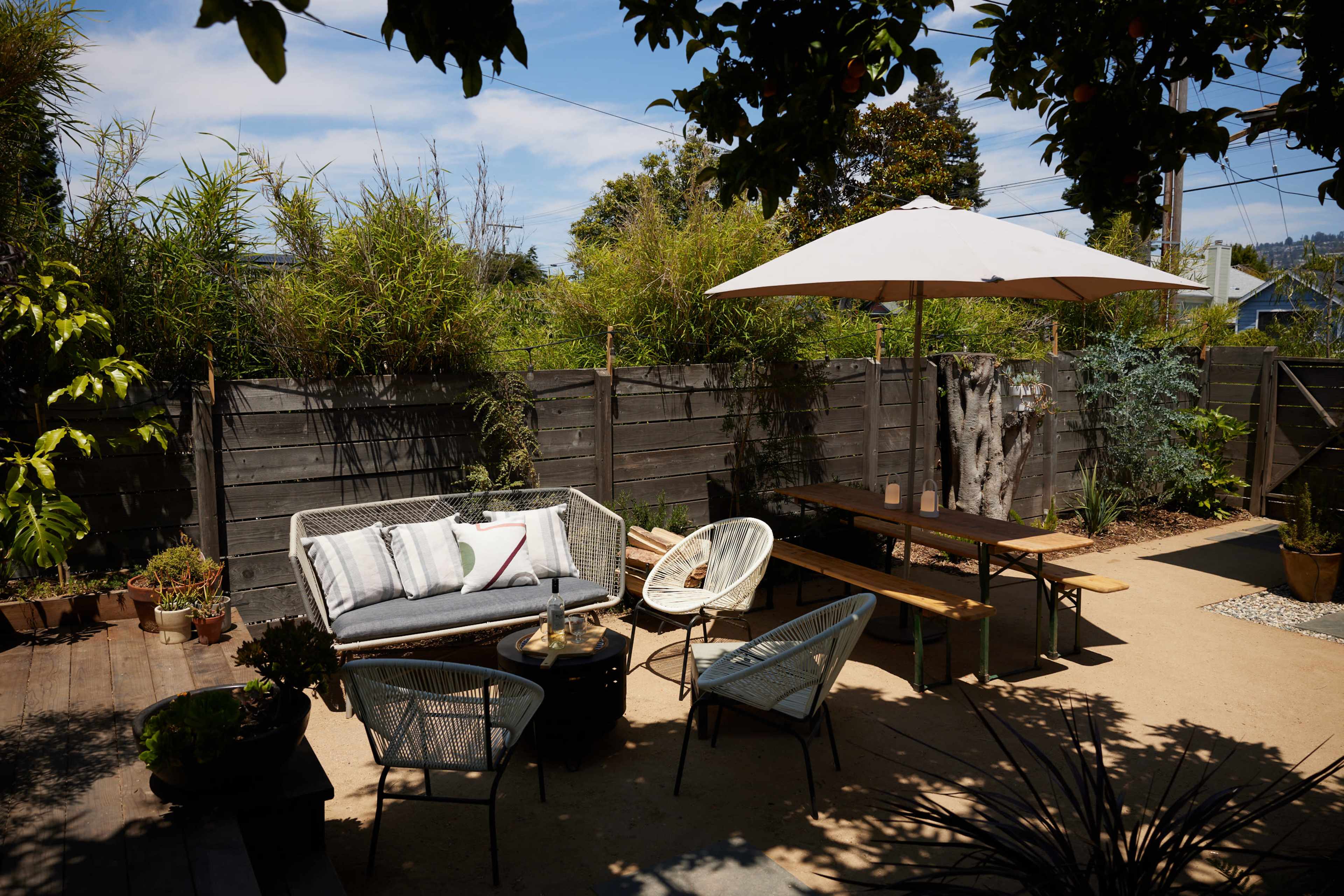 The image shows an outdoor patio area with a sofa, chairs, a table, and an umbrella, surrounded by plants and a wooden fence.