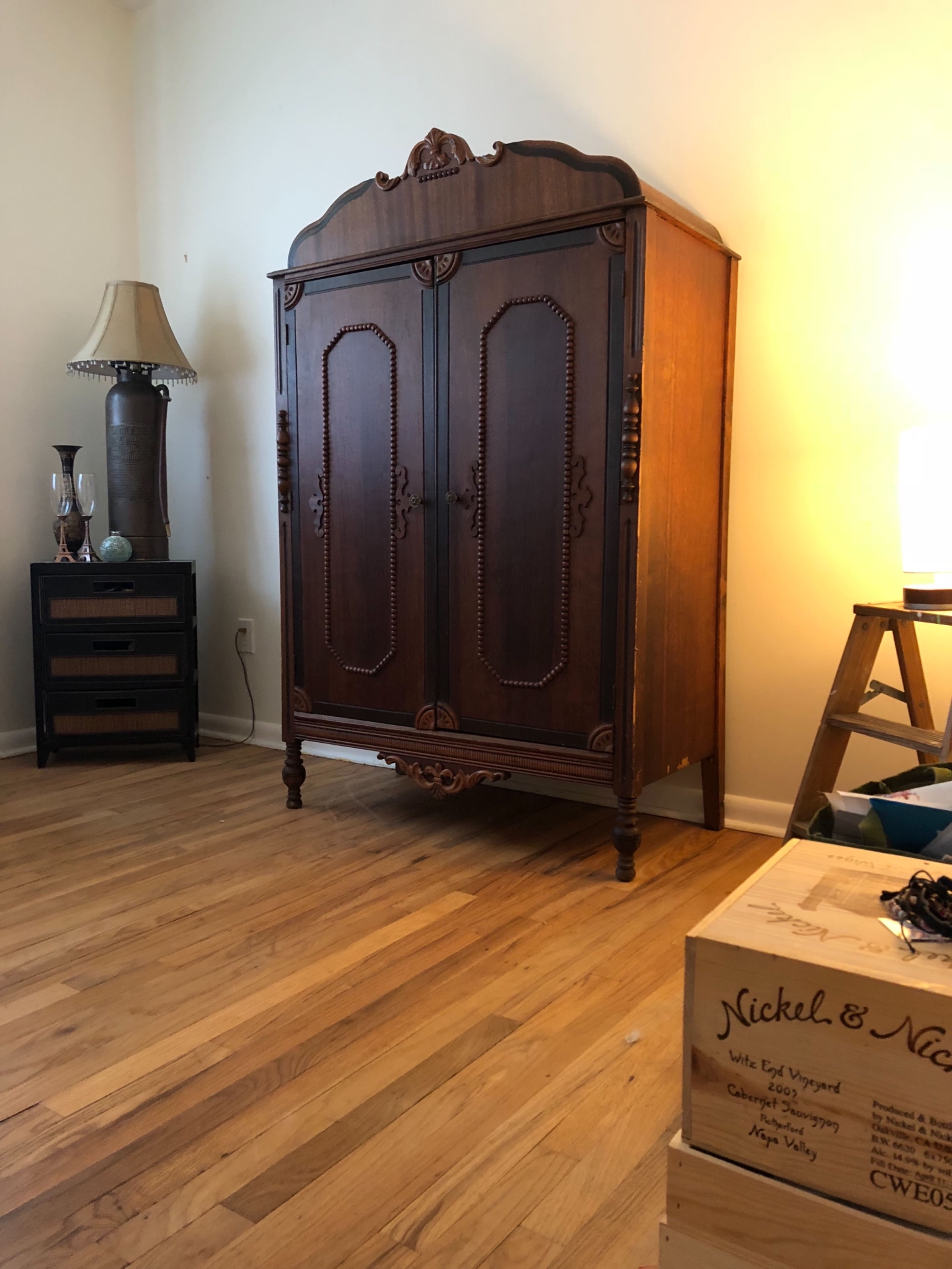 The image shows a wooden armoire with ornate carvings, placed against a wall in a room with hardwood flooring, alongside a lamp and a nightstand.