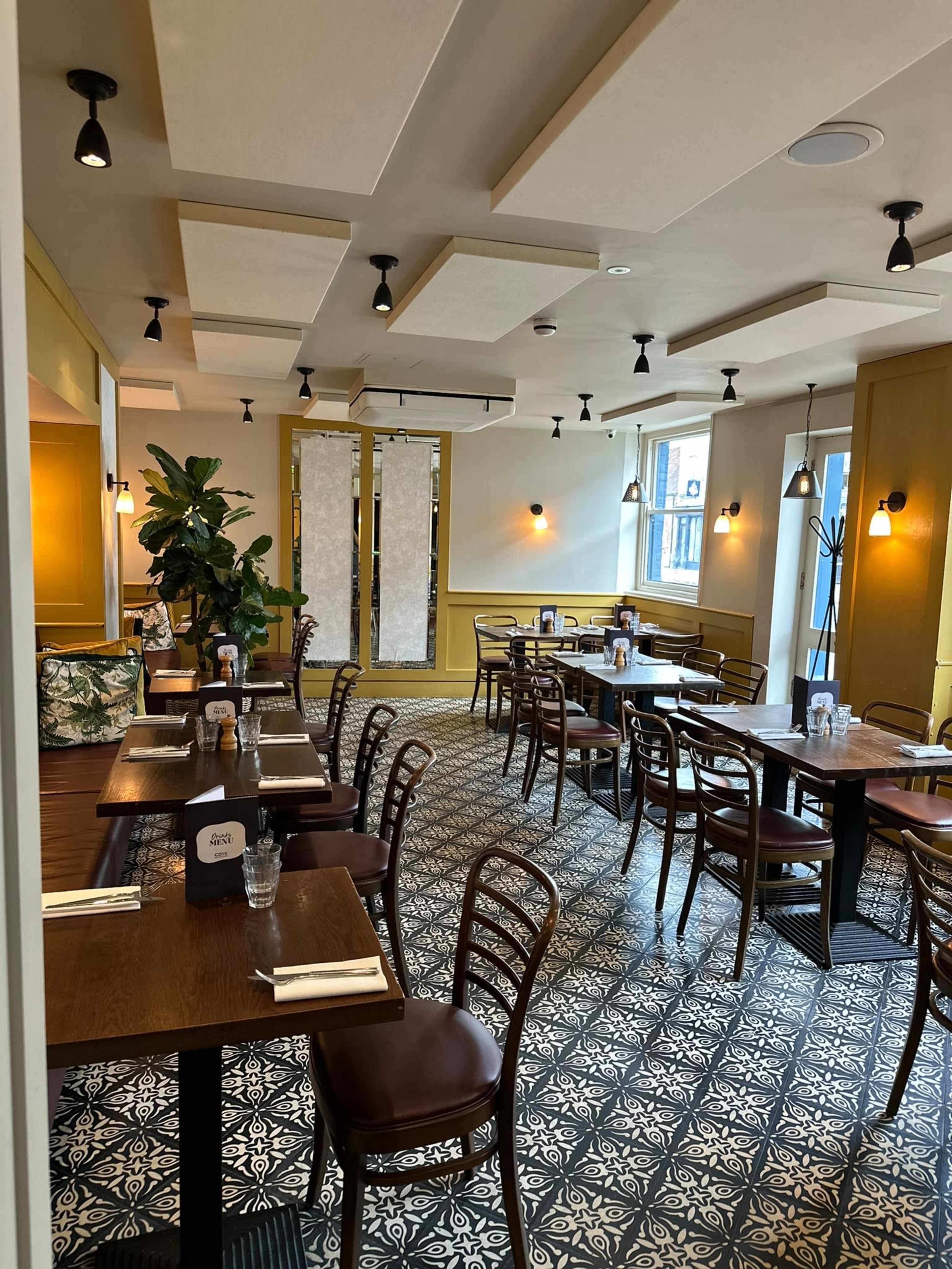 The image shows a well-lit restaurant interior with wooden tables and chairs arranged neatly on a patterned tiled floor, featuring plants and decorative wall elements.