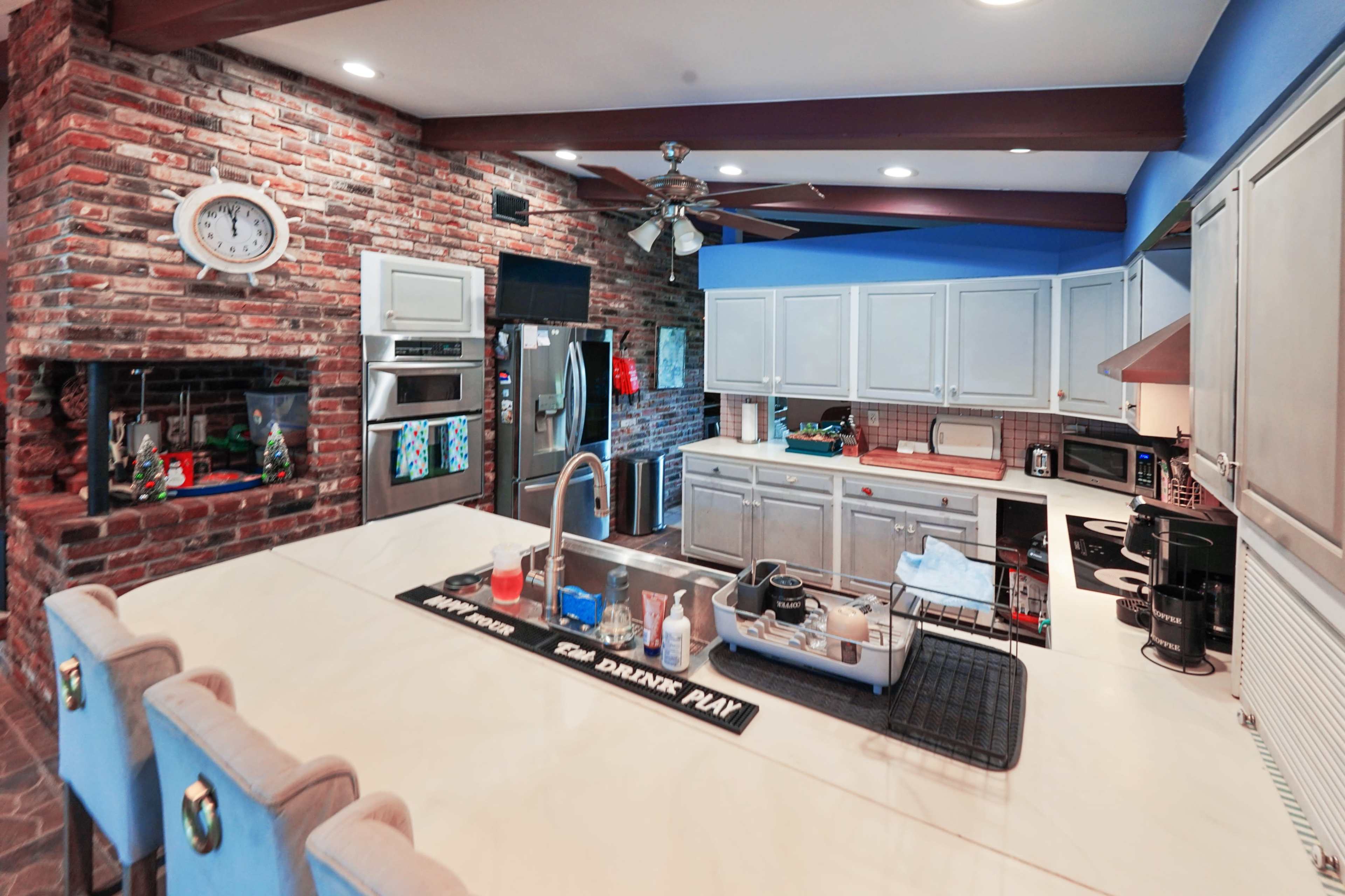 The image shows a spacious kitchen with brick walls, a large island with a sink, stainless steel appliances, and a ceiling fan.