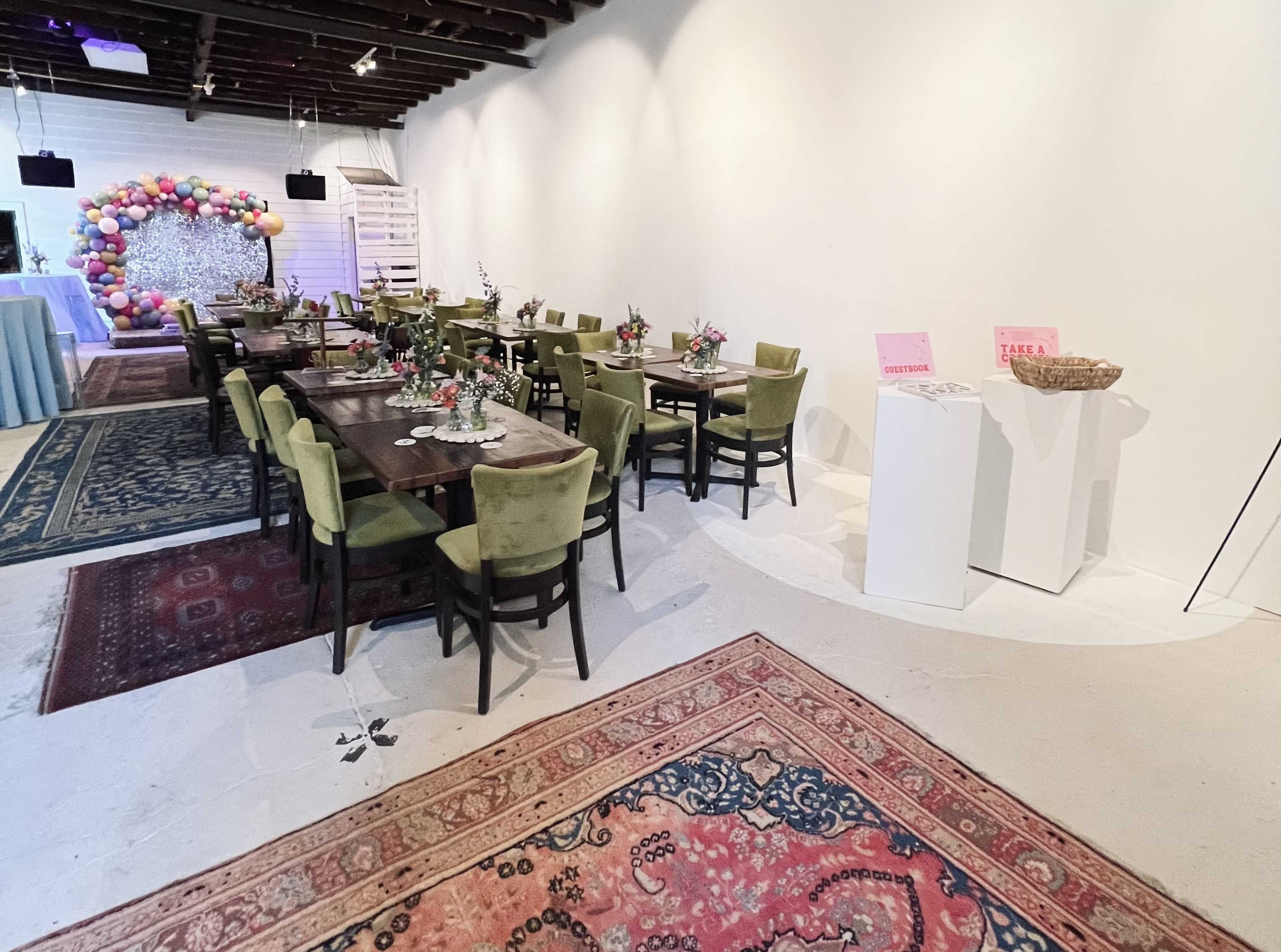 A spacious event room with several dining tables surrounded by green chairs, decorative floral arrangements, and a backdrop of colorful balloons.
