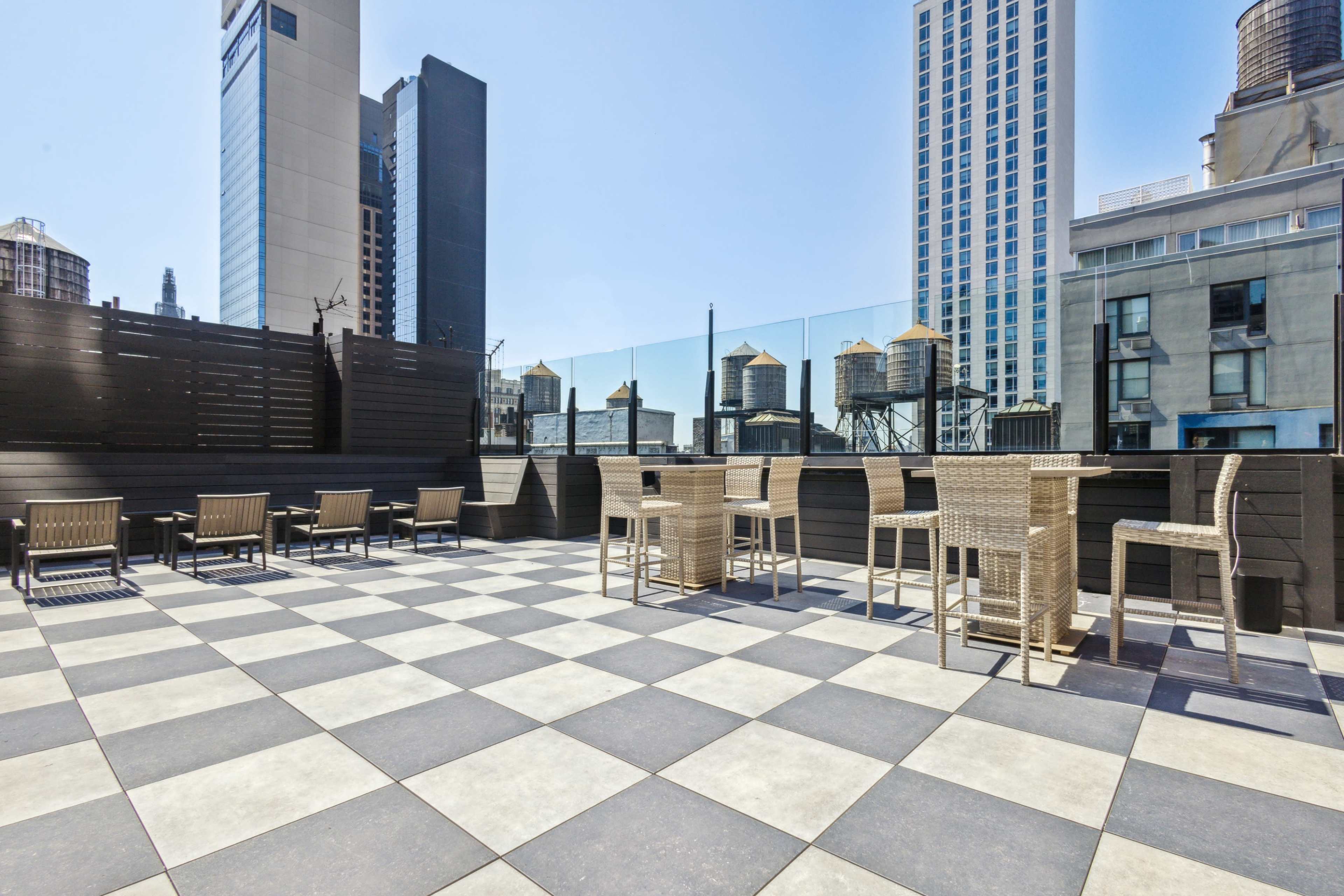 A rooftop terrace with checkered flooring, featuring tall wicker bar stools and tables, overlooking buildings with water towers in the background.