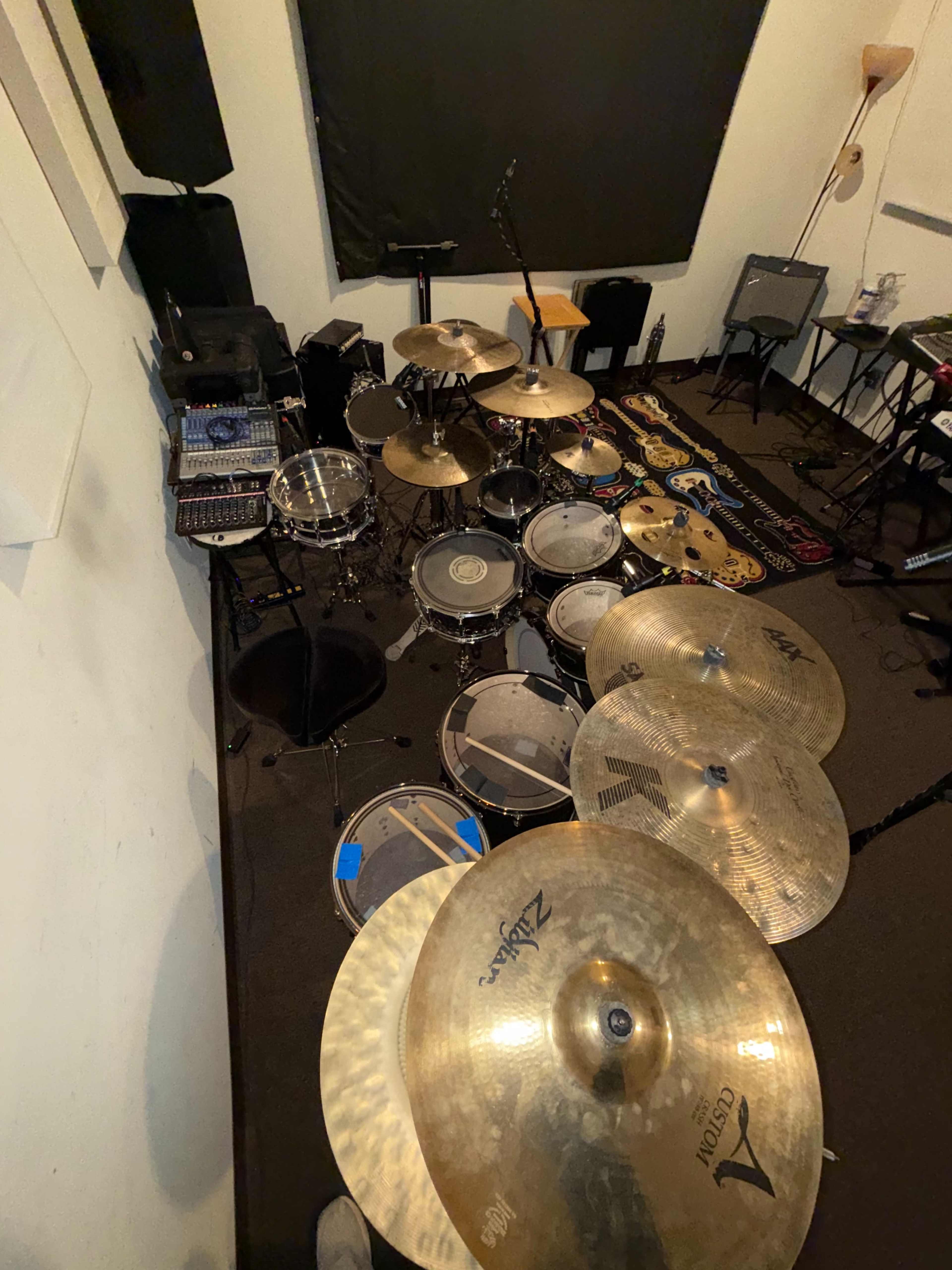 The image shows a rehearsal space with a large drum set, various percussion instruments, electronic equipment, and a microphone setup.