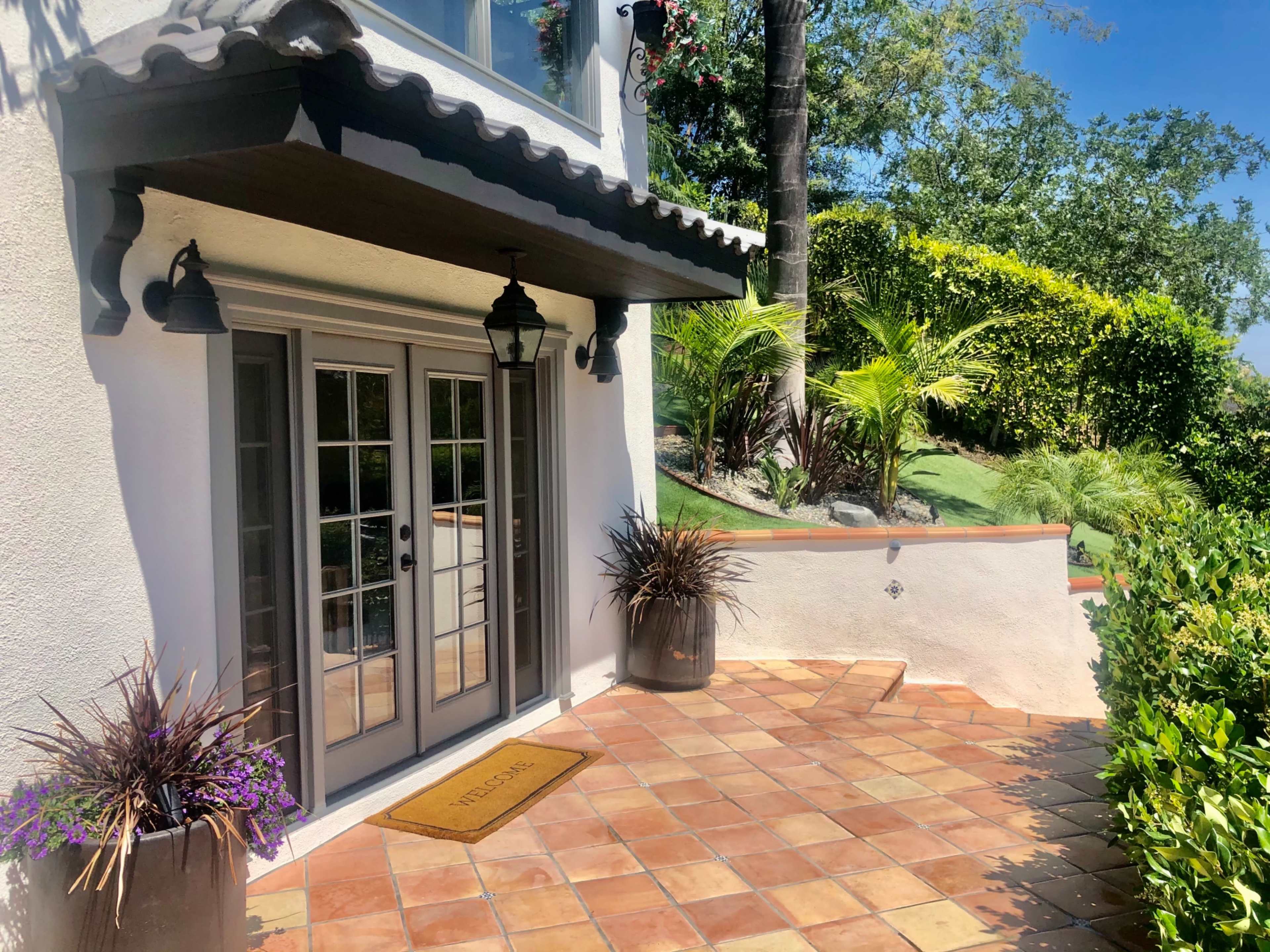 The image shows a home's entrance featuring double doors, a welcome mat, and potted plants on a tiled patio surrounded by greenery.
