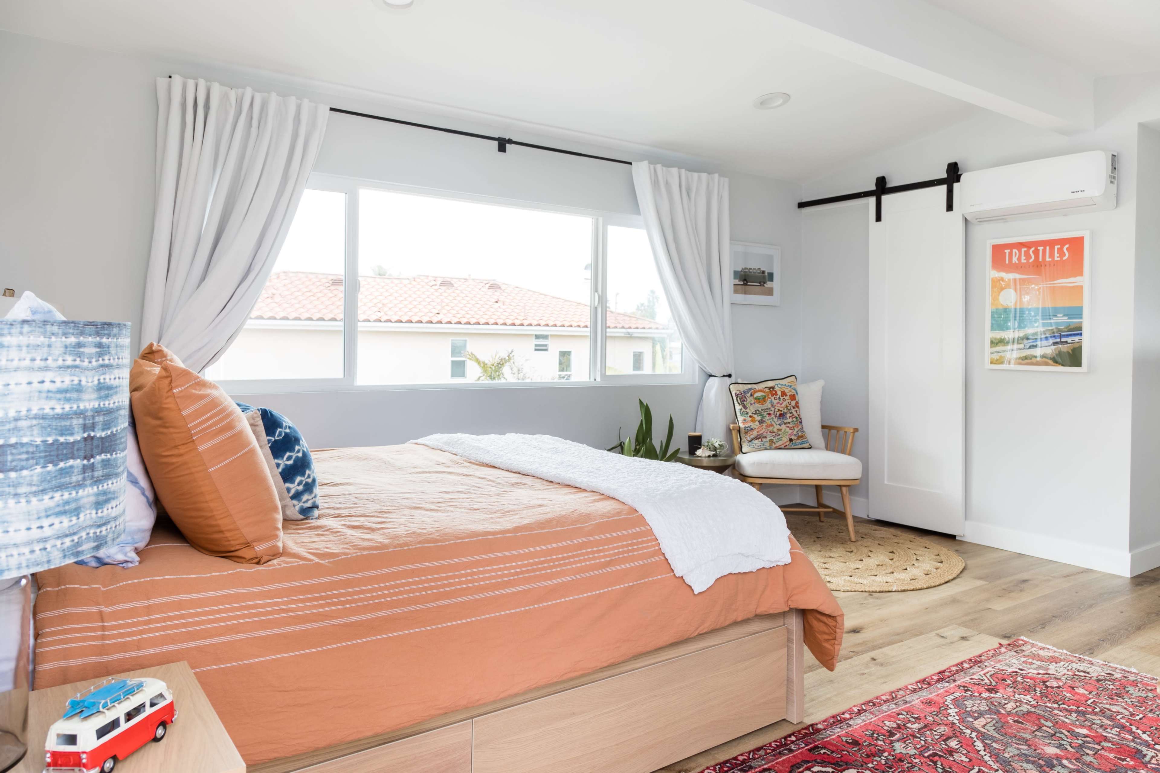 The image features a bright bedroom with a bed covered in an orange duvet, a small decorative chair, and large windows allowing natural light to fill the space.