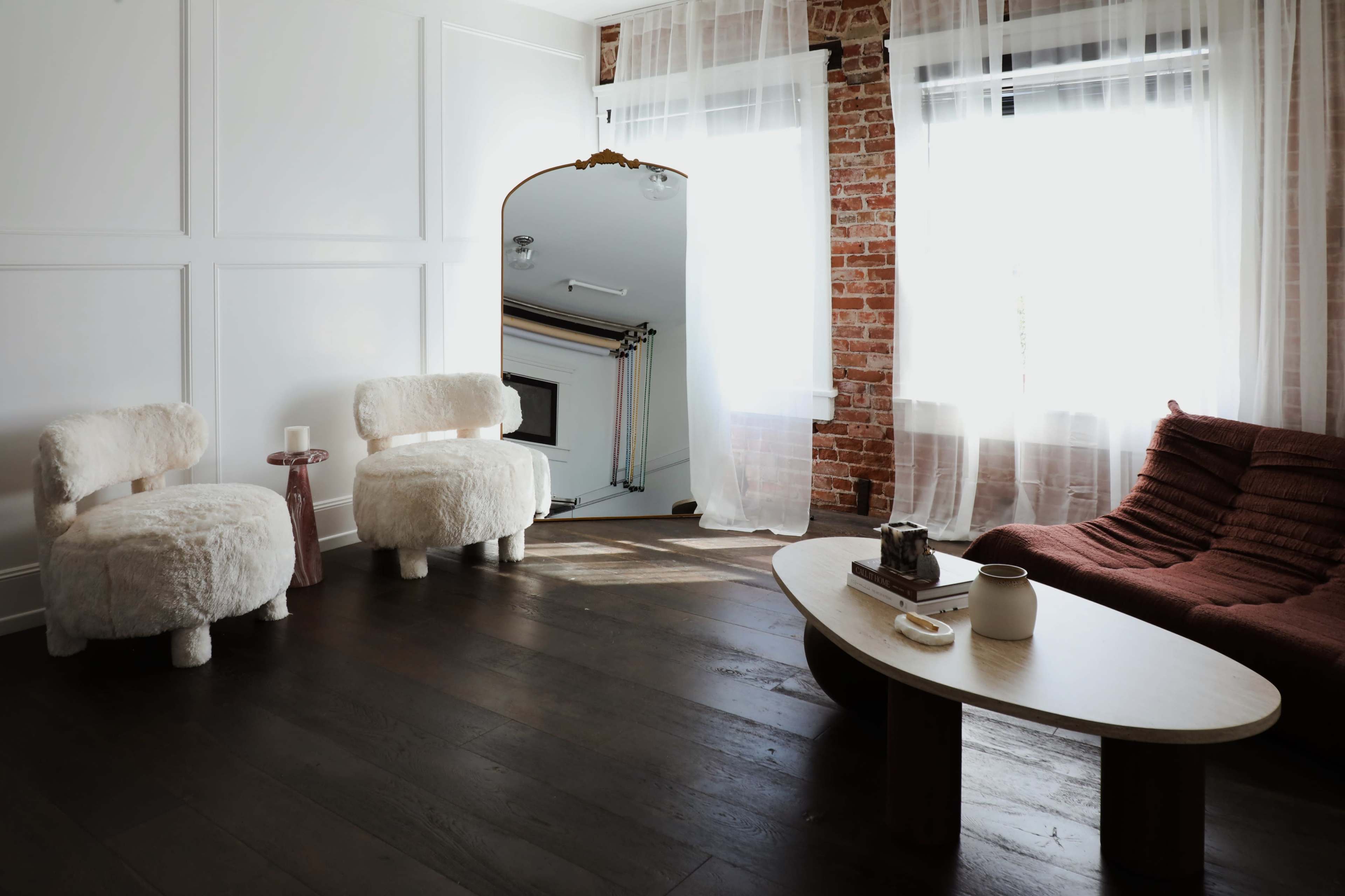 The room features a pair of furry white chairs, a red textured sofa, a wooden coffee table, and a large mirror against a backdrop of exposed brick walls and light drapes.