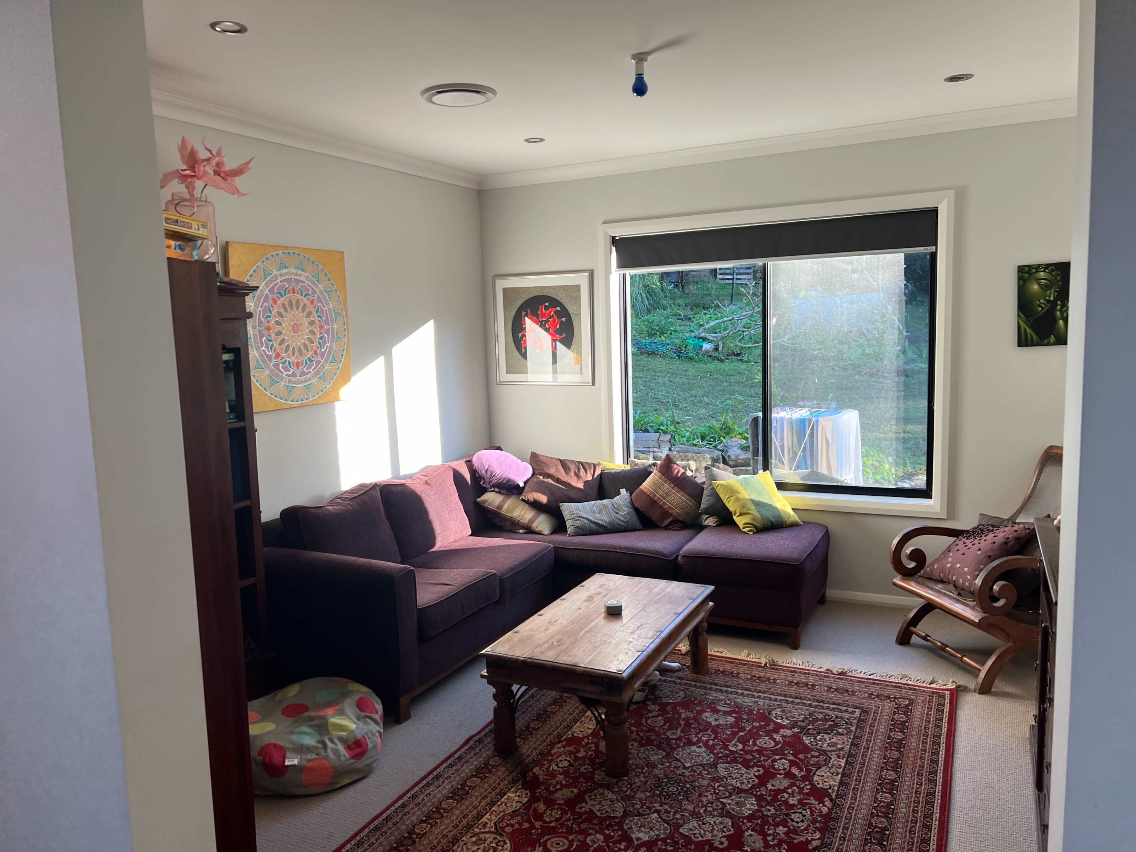 A cozy living room features a purple sectional sofa, a wooden coffee table, and large windows overlooking a green yard.