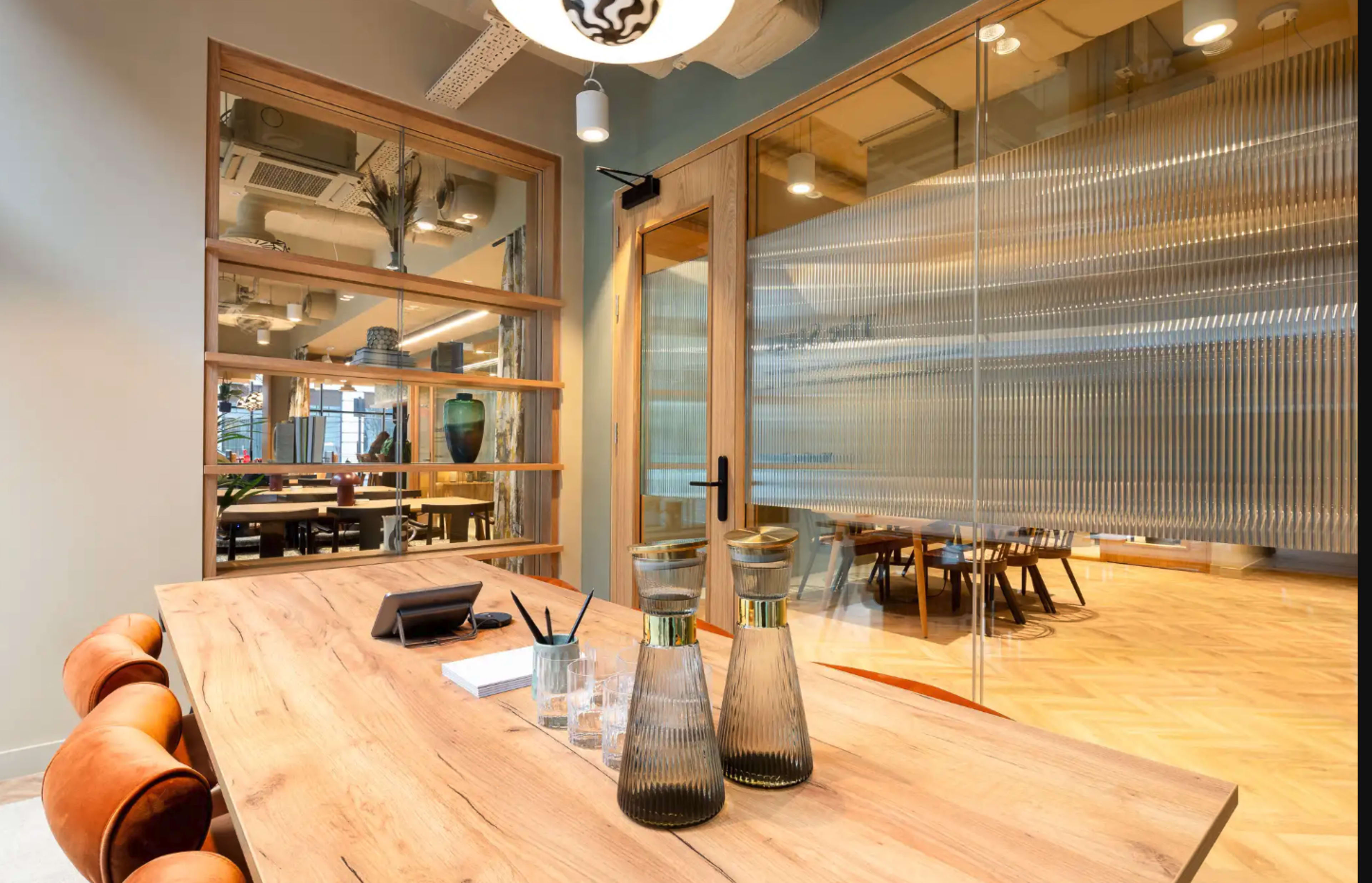 A wooden table with glass vases sits in a bright office space, featuring a glass-walled meeting room in the background.