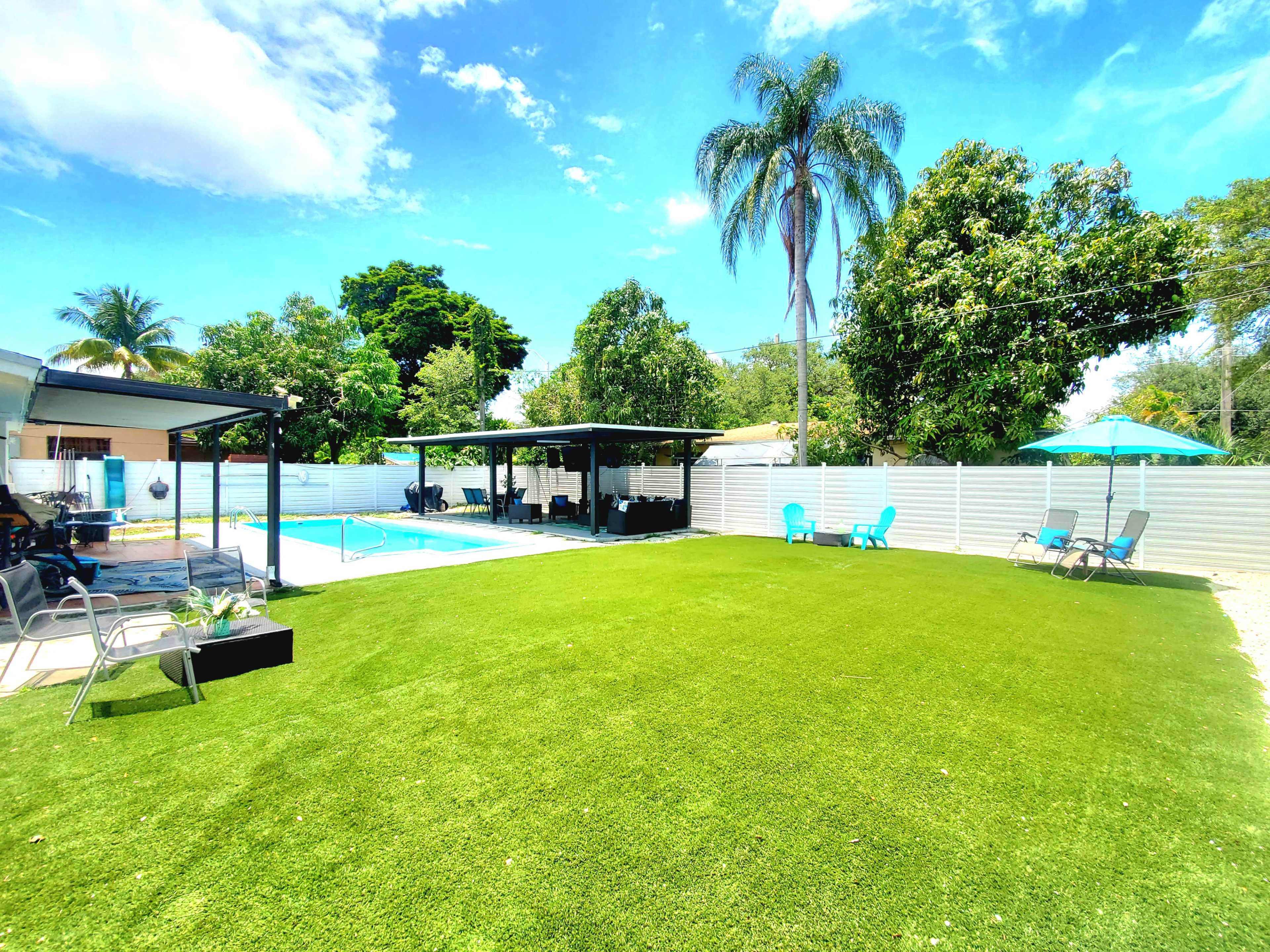 The image shows a backyard with a swimming pool, a patio area with seating, and artificial grass surrounded by trees and a white fence under a blue sky.