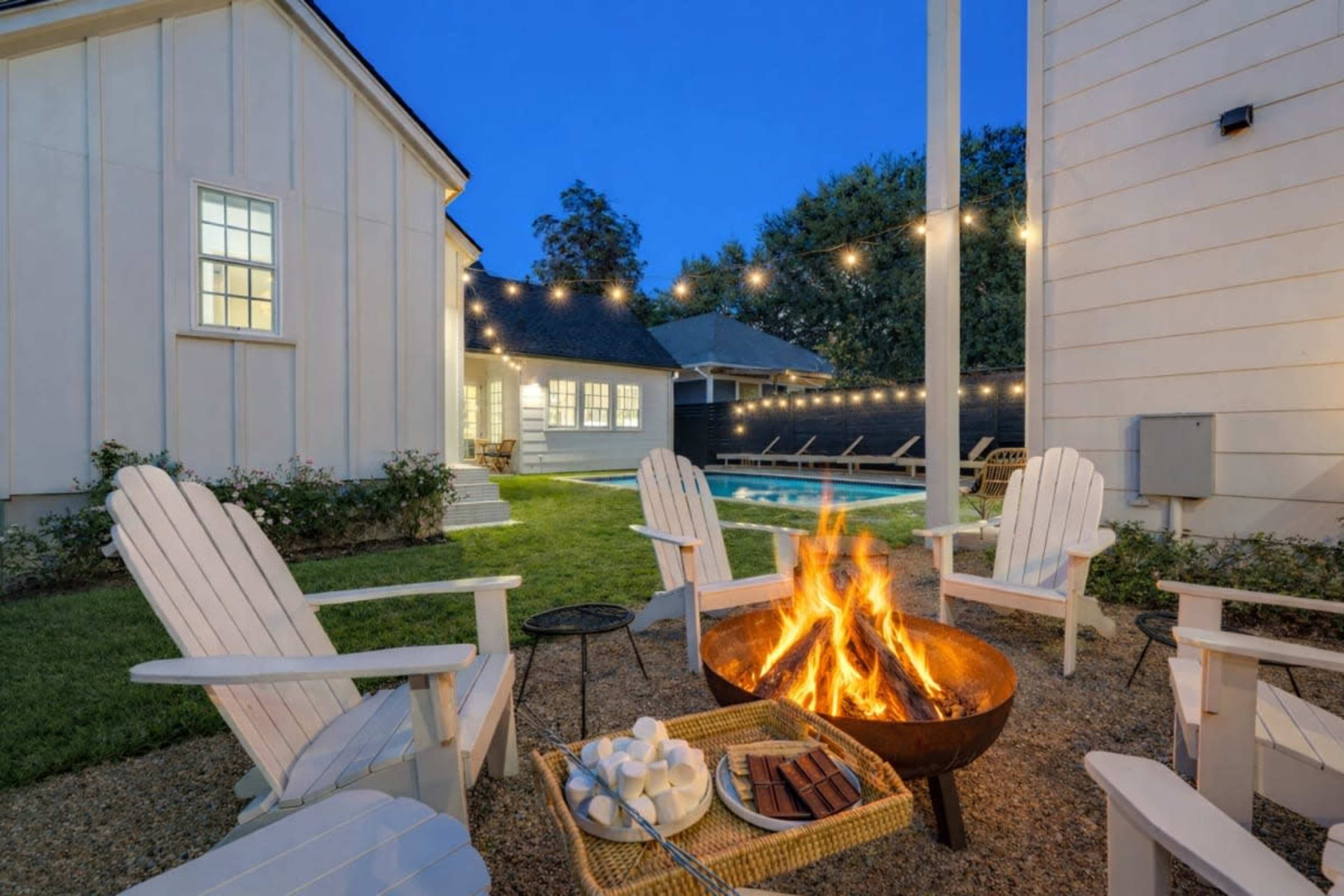 A cozy outdoor seating area features four white adirondack chairs arranged around a fire pit, with a pool and string lights visible in the background.
