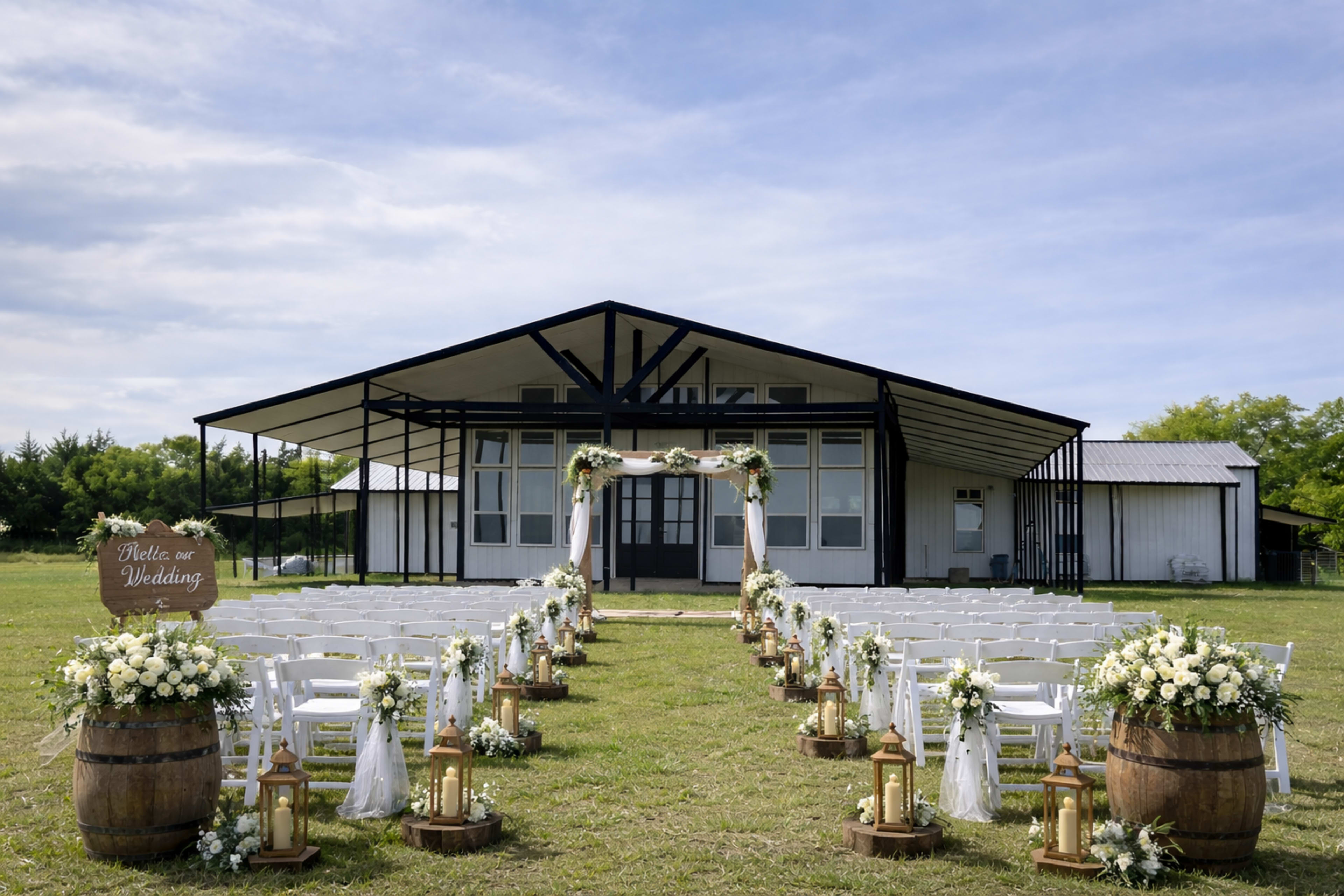 A rustic wedding venue features a large, modern building with rows of white chairs set up along a flower-adorned aisle leading to an archway.