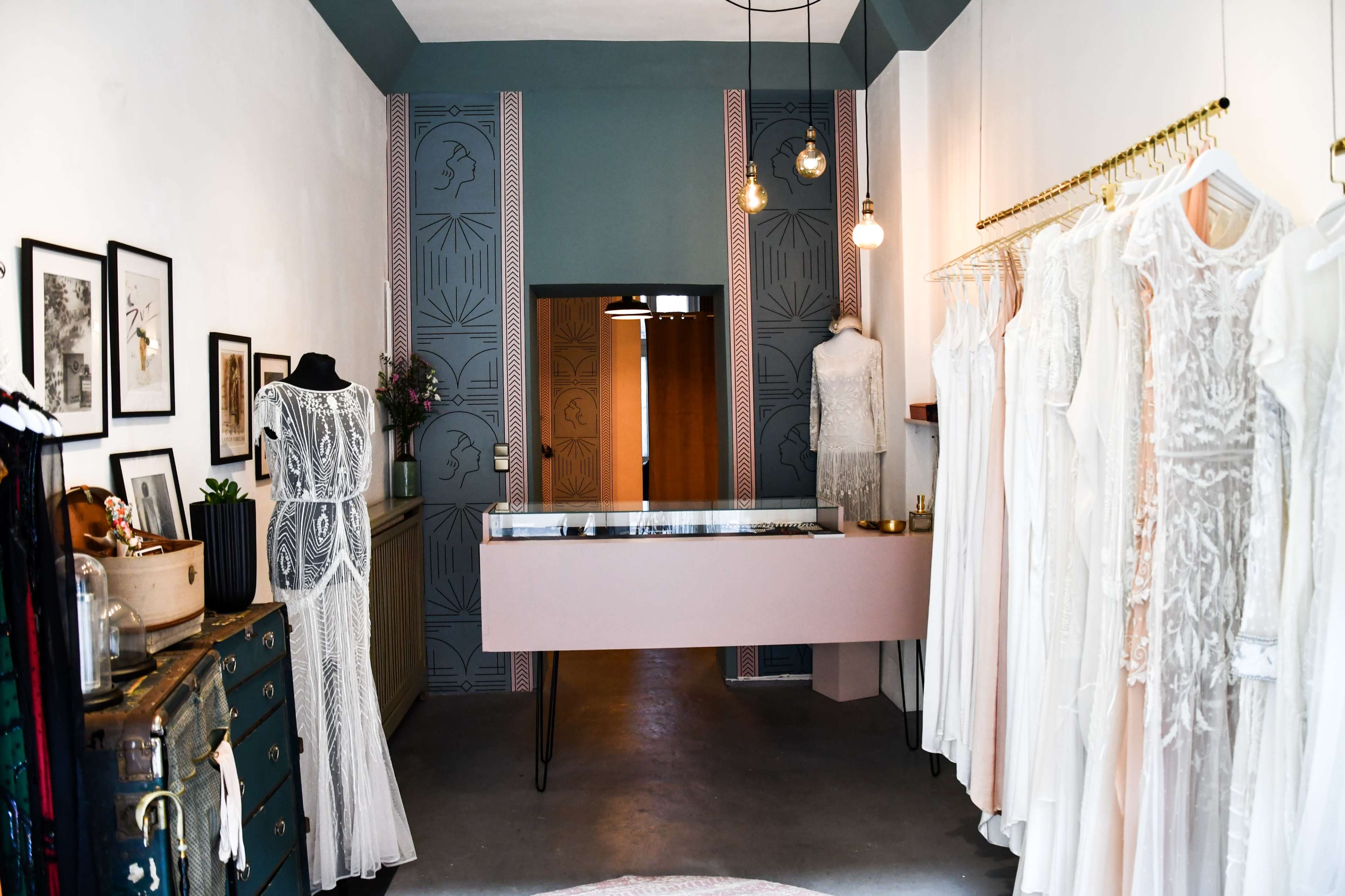 The image shows a modern boutique displaying elegant dresses, with a pale pink display counter, adorned walls, and various decor elements.