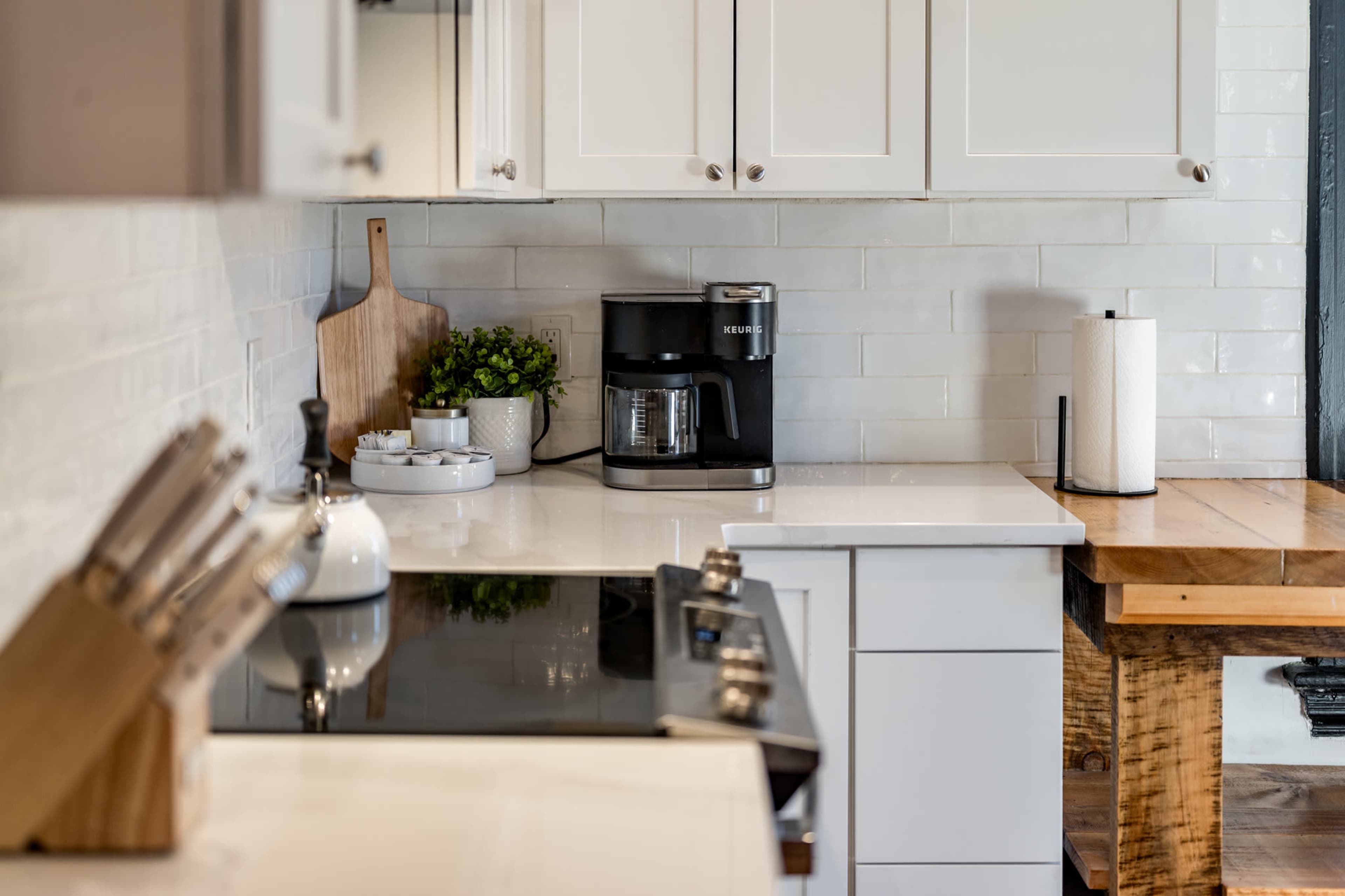 A modern kitchen features a black coffee maker on a countertop with white cabinetry and a wooden table.