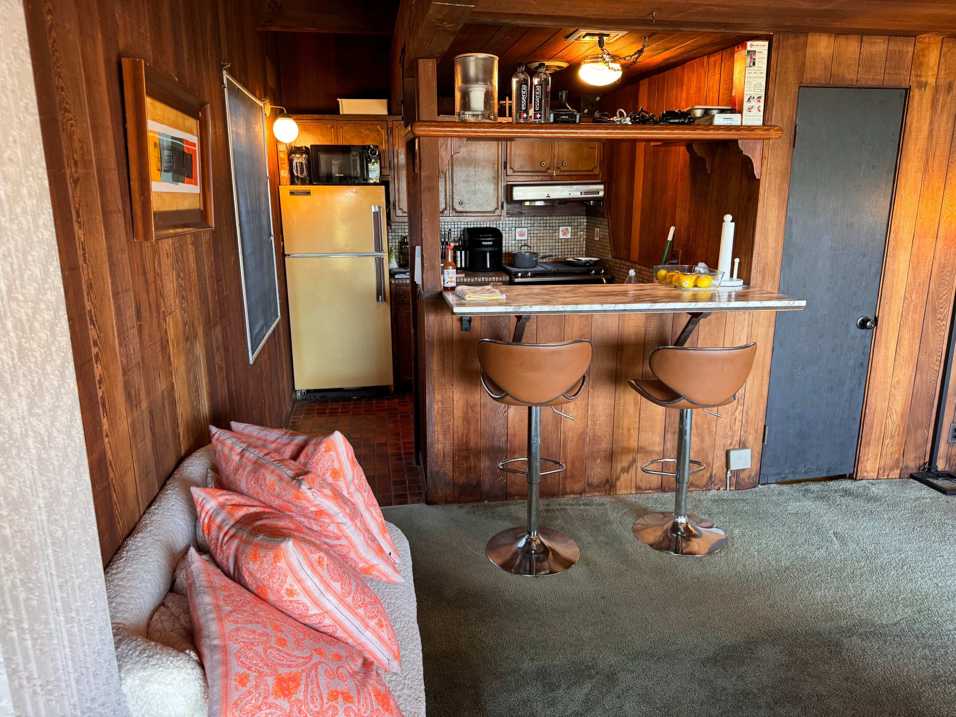 A small, wooden interior with a kitchen bar featuring two stools, a sofa with patterned pillows, and a view of a refrigerator and cooking area.