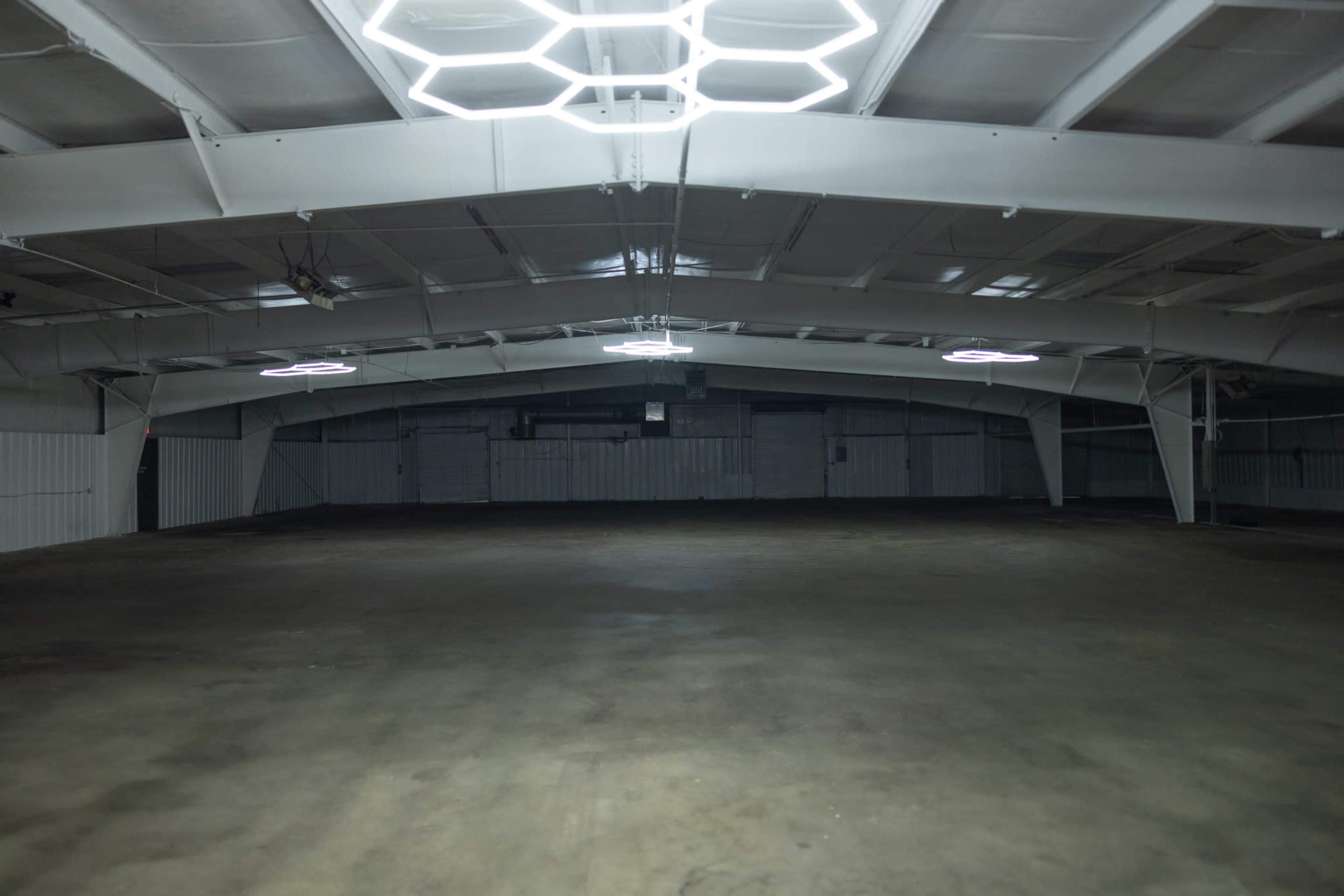 A spacious and empty warehouse features exposed steel beams and hexagonal light fixtures hanging from the ceiling.