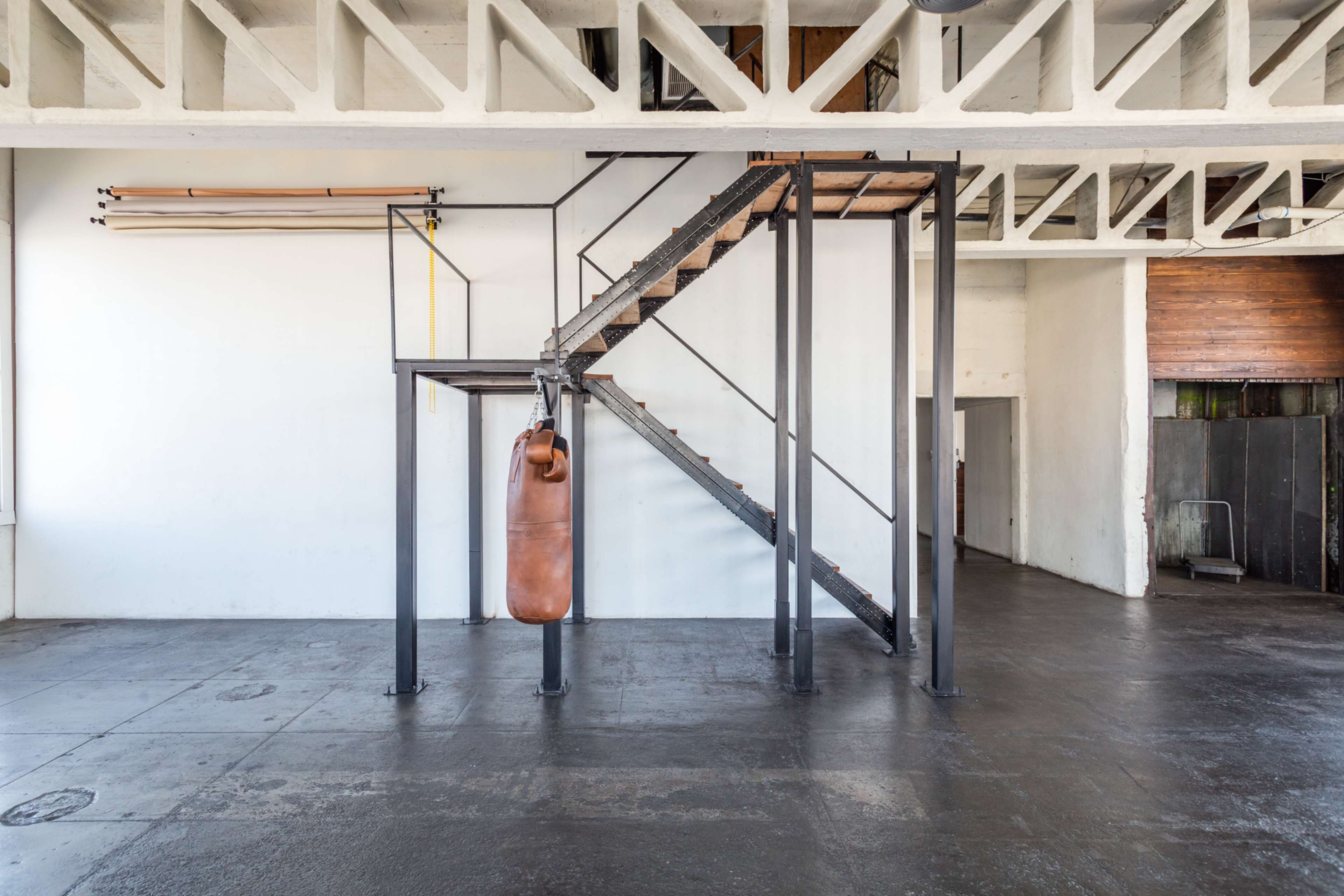 A spacious, industrial-style gym featuring a brown leather punching bag hanging beside a staircase with metal supports and wooden steps.