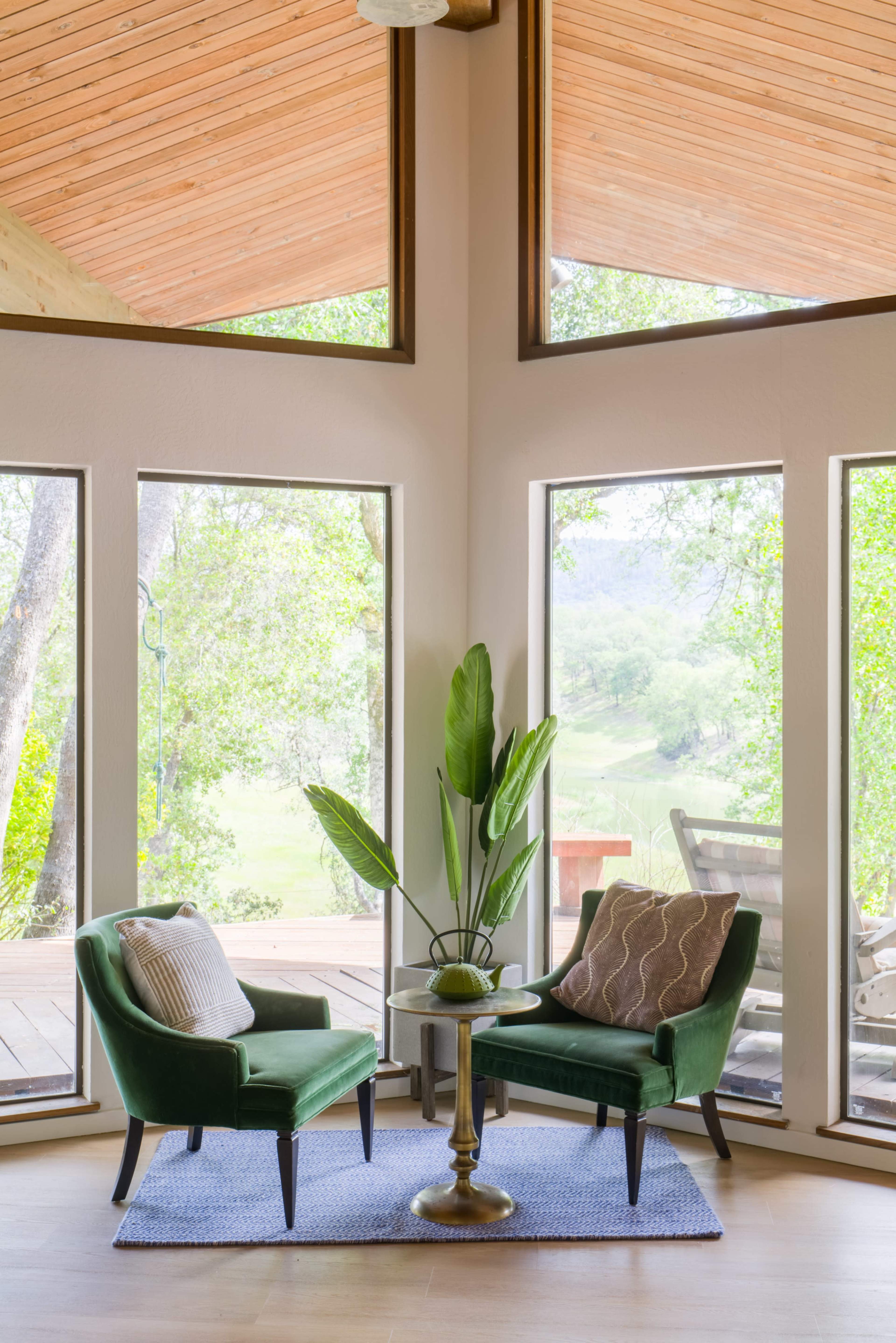 A bright corner of a room features two green chairs and a circular table, facing large windows that offer a view of the outdoors.