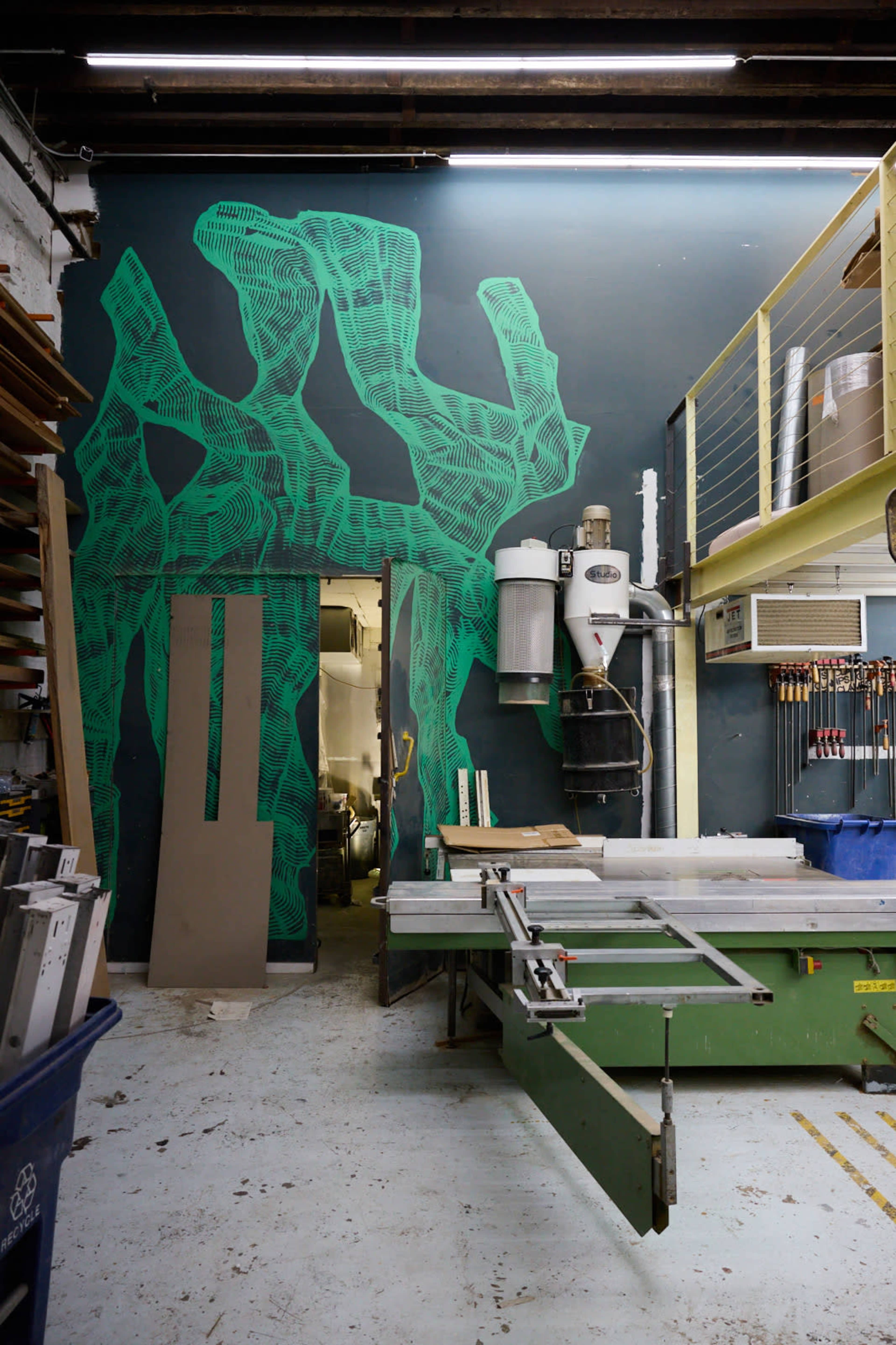 The image shows a workshop with a large green abstract mural on the wall, featuring tools and equipment, including a saw and a dust collection system.