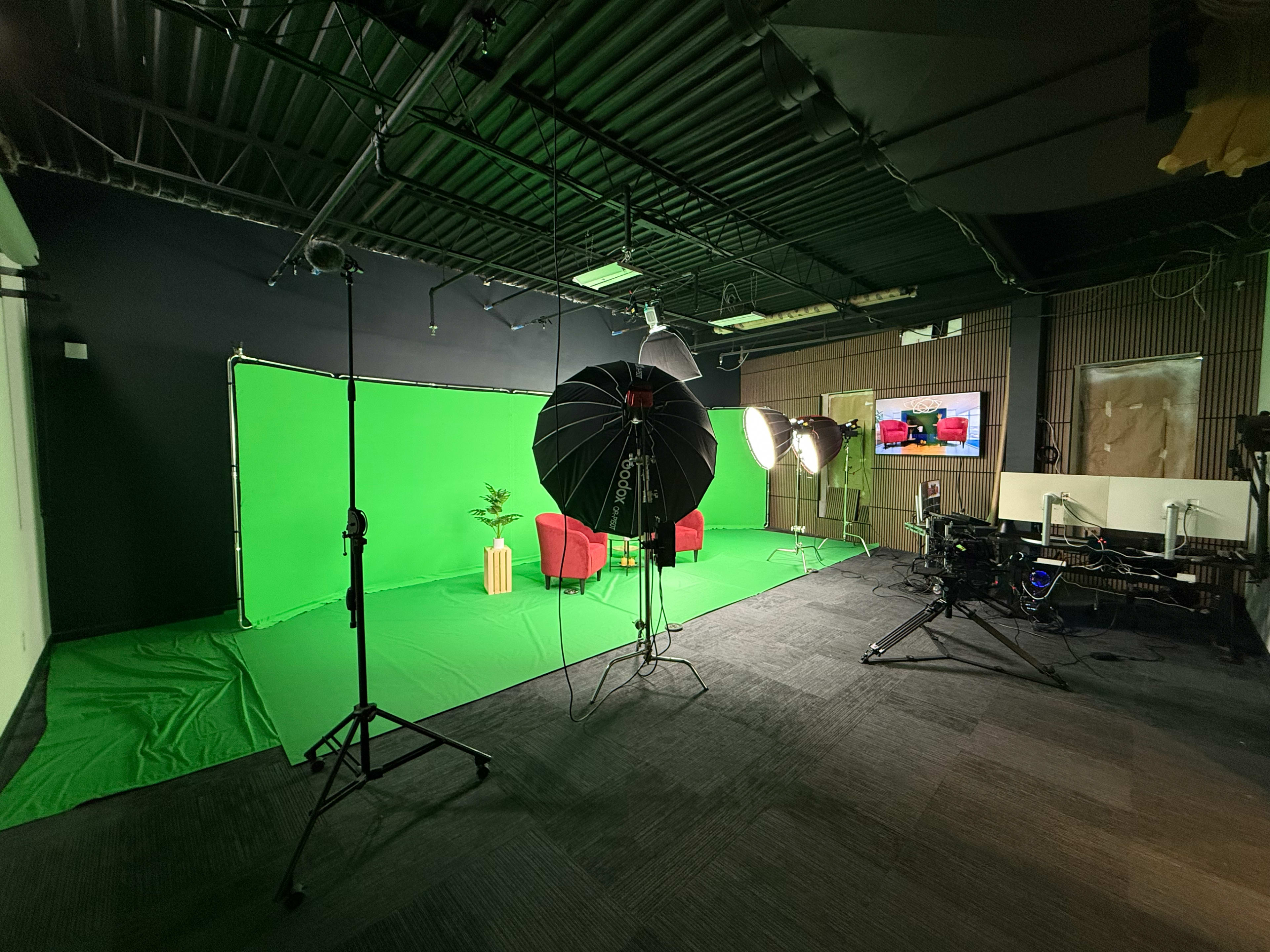 The image shows a studio set up with a green screen, two red chairs, lighting equipment, and a camera on a tripod.