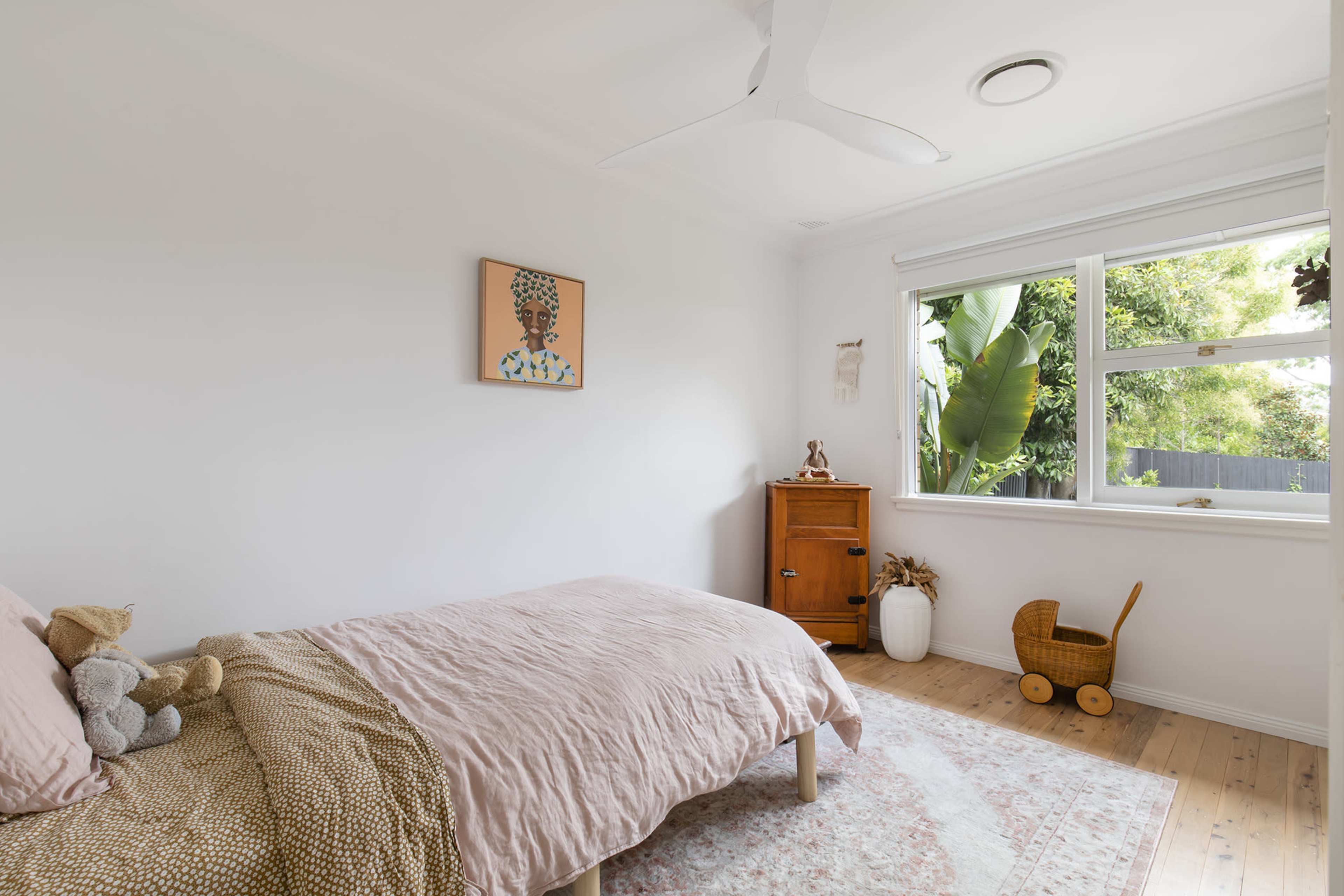A simple bedroom features a bed with pink bedding, a wooden cabinet, a soft rug, and a window overlooking greenery.