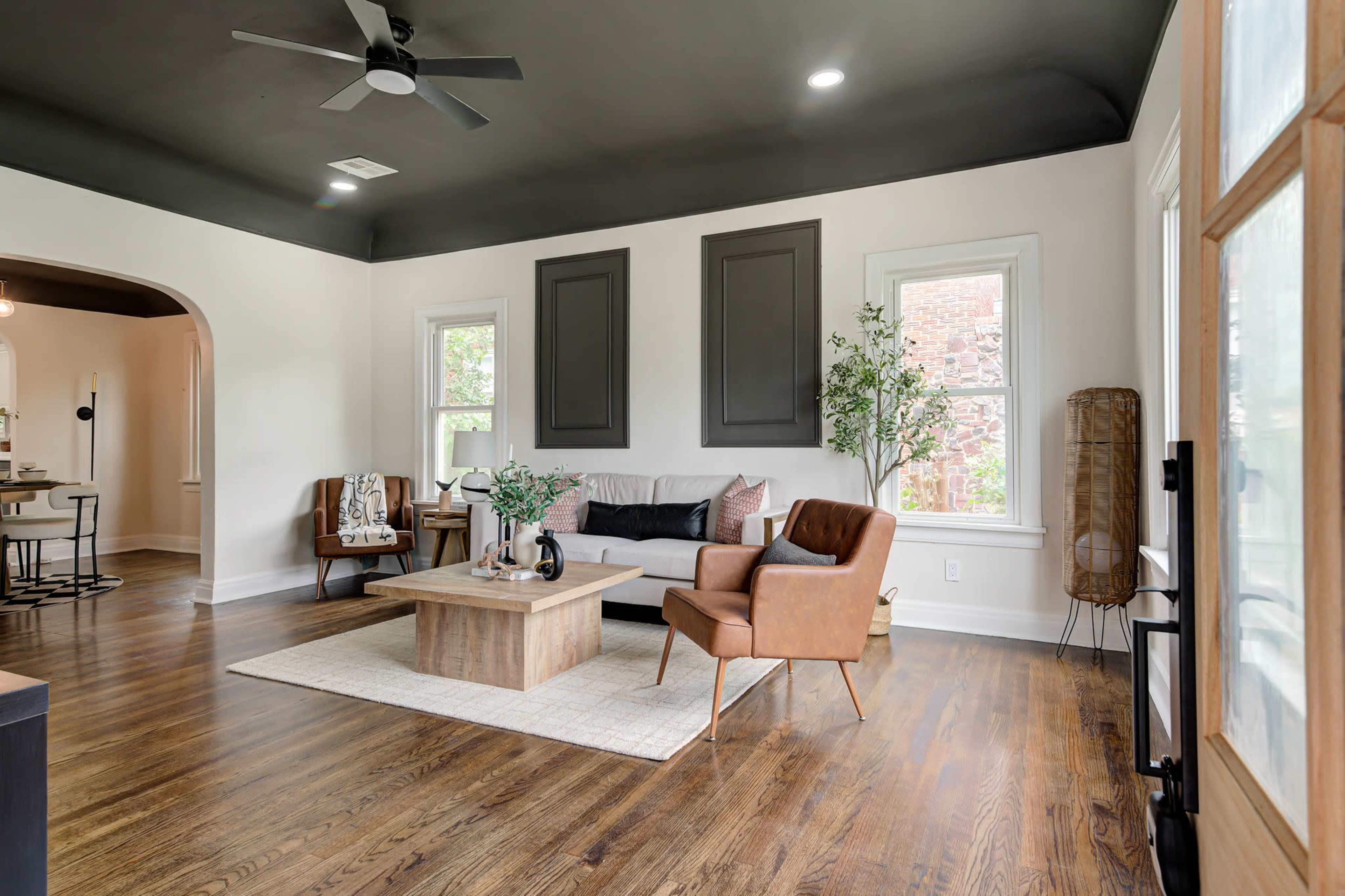 The image shows a spacious living room with a dark ceiling, a comfortable sofa, two armchairs, a coffee table, and large windows allowing natural light.