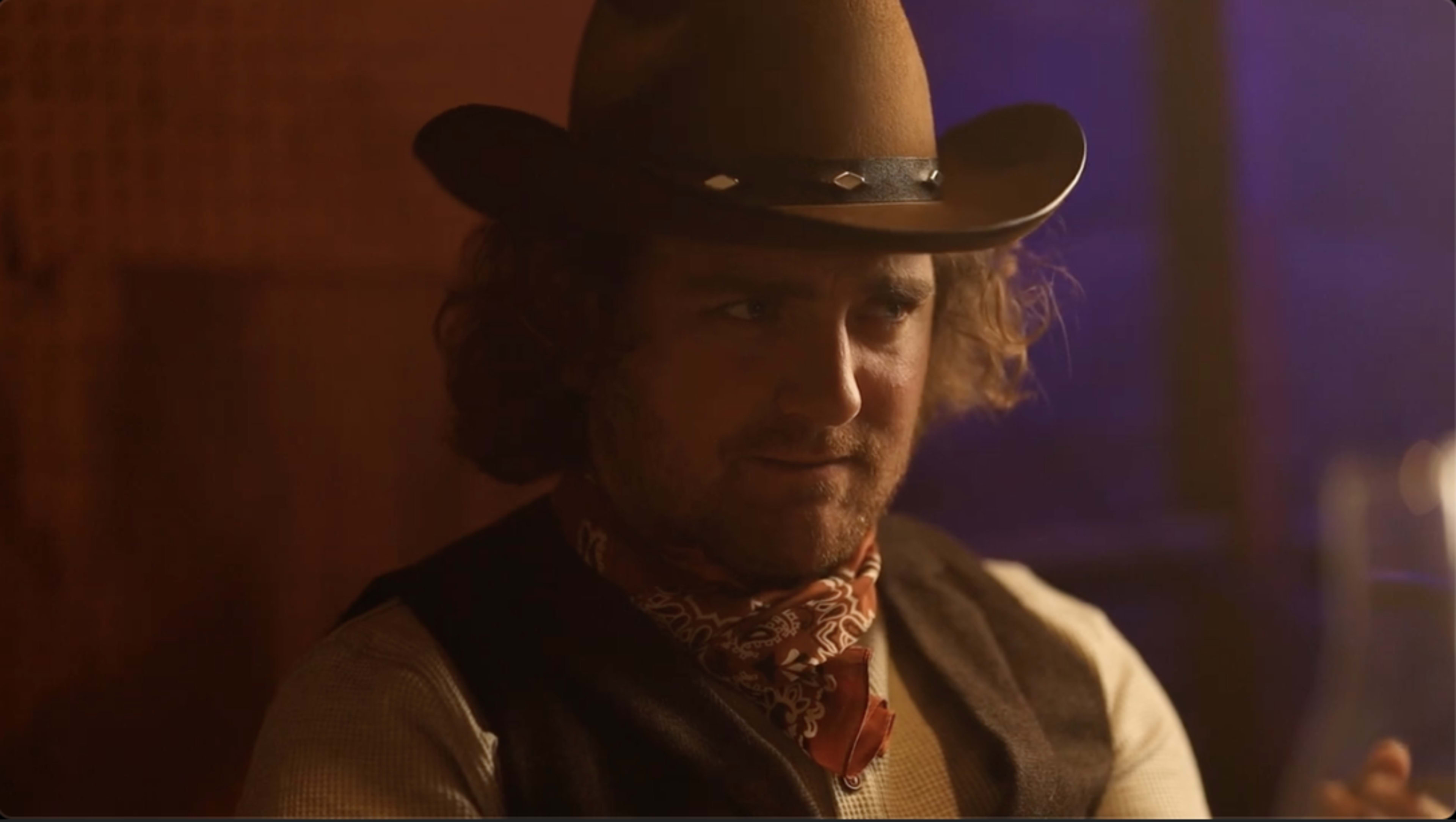 A man wearing a cowboy hat and a bandana sits at a table, looking intently at a glass.