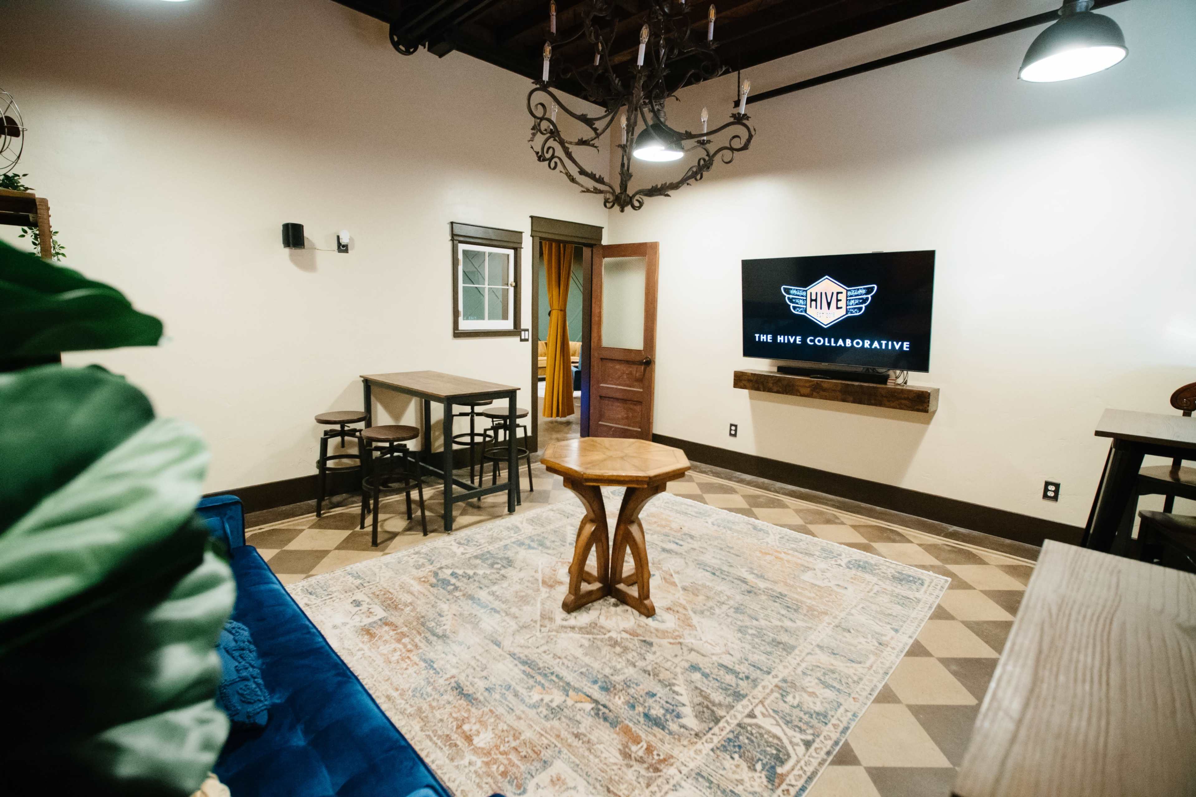 A cozy lounge area with a blue sofa, a wooden coffee table in the center, a large TV displaying the logo "HIVE COLLABORATIVE," and a decorative rug on a tiled floor.