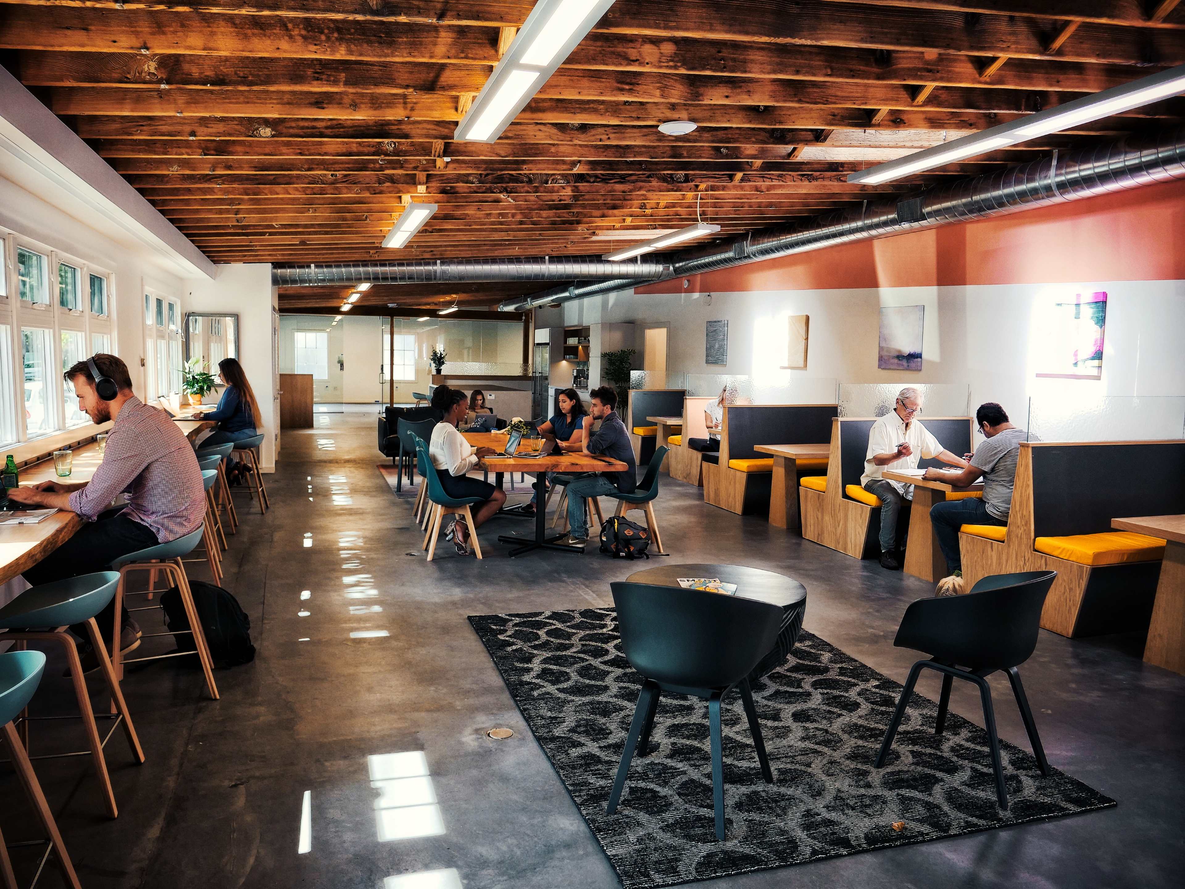 A modern open office space with wooden beams, various seating areas, and people engaged in work and conversation.