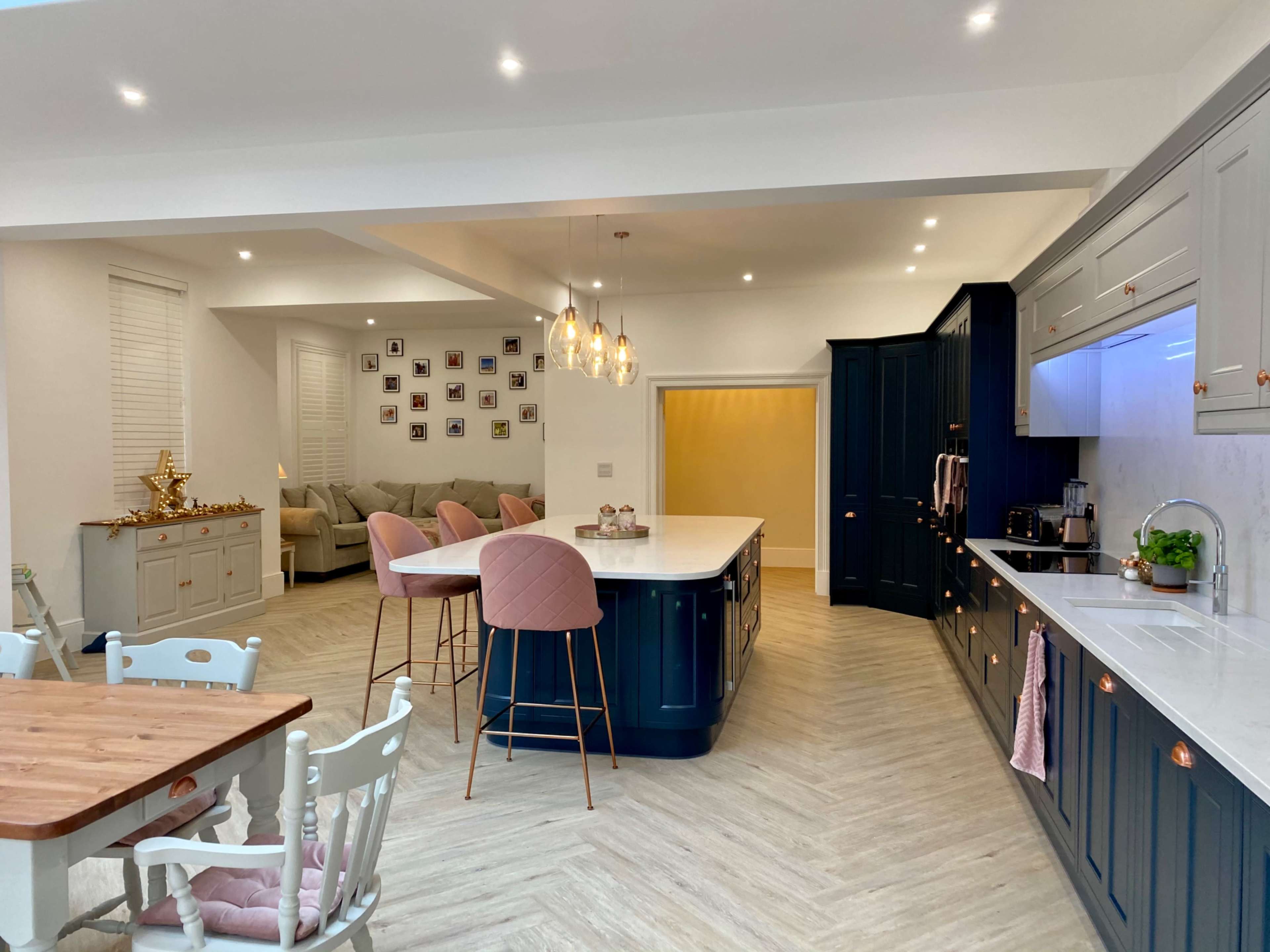 The image shows a modern kitchen with a central island, pink bar stools, and light wood flooring, adjacent to a living area with a wall of photographs.