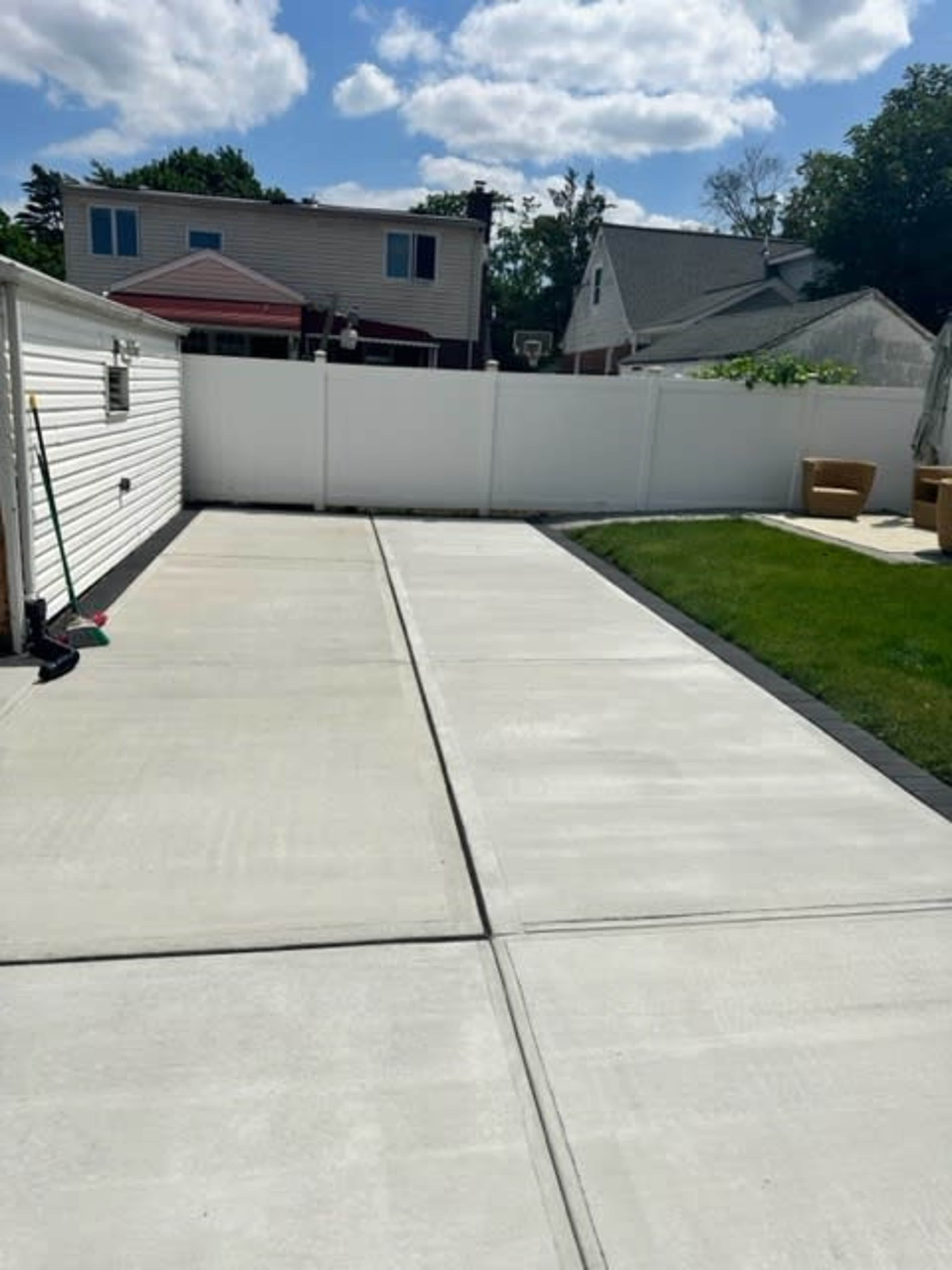 A concrete driveway or patio area beside a green lawn, enclosed by a white fence with residential houses in the background.