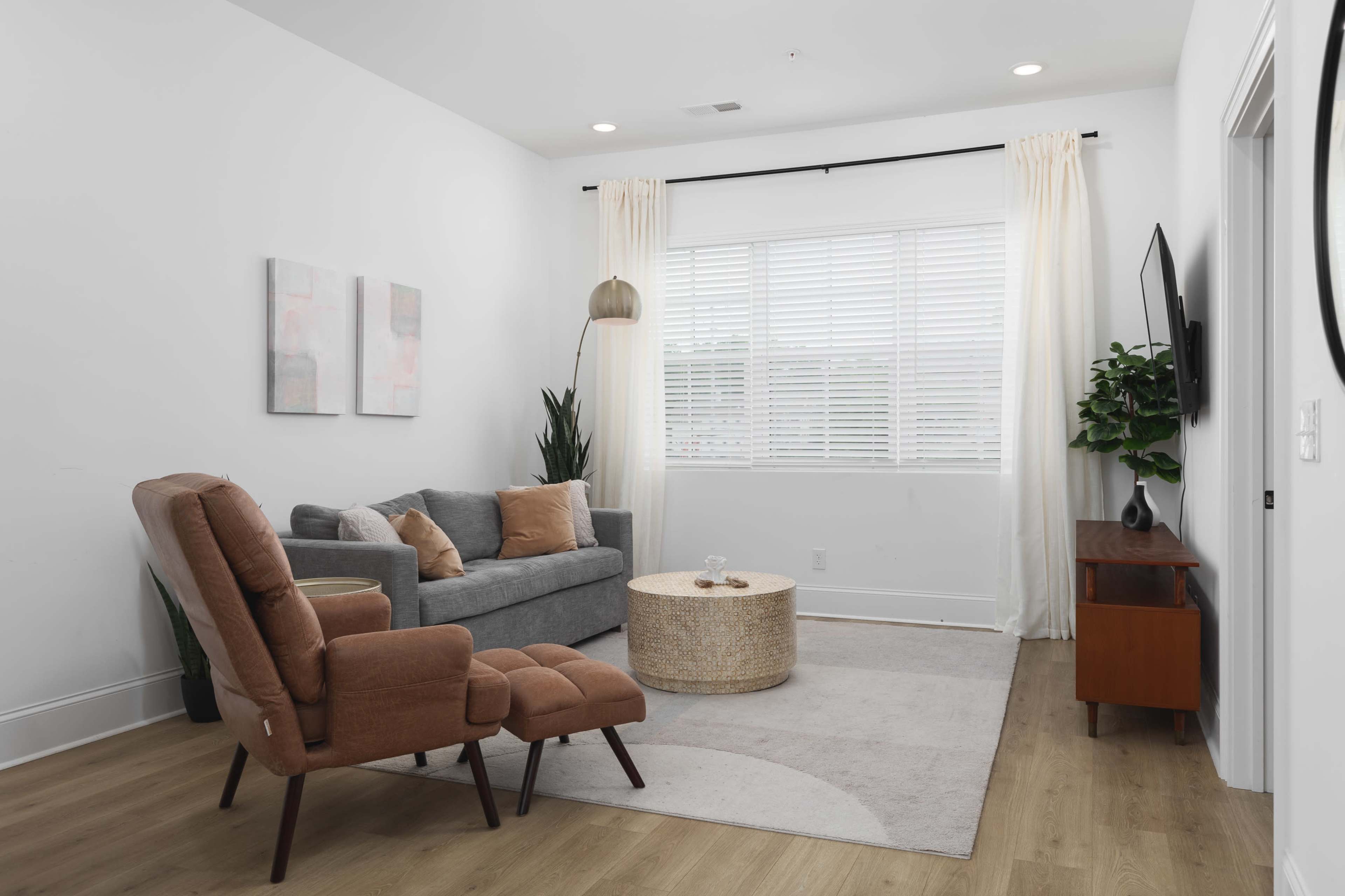 The room features a gray sofa, an armchair, a round table, a television on the wall, and large windows with white blinds.