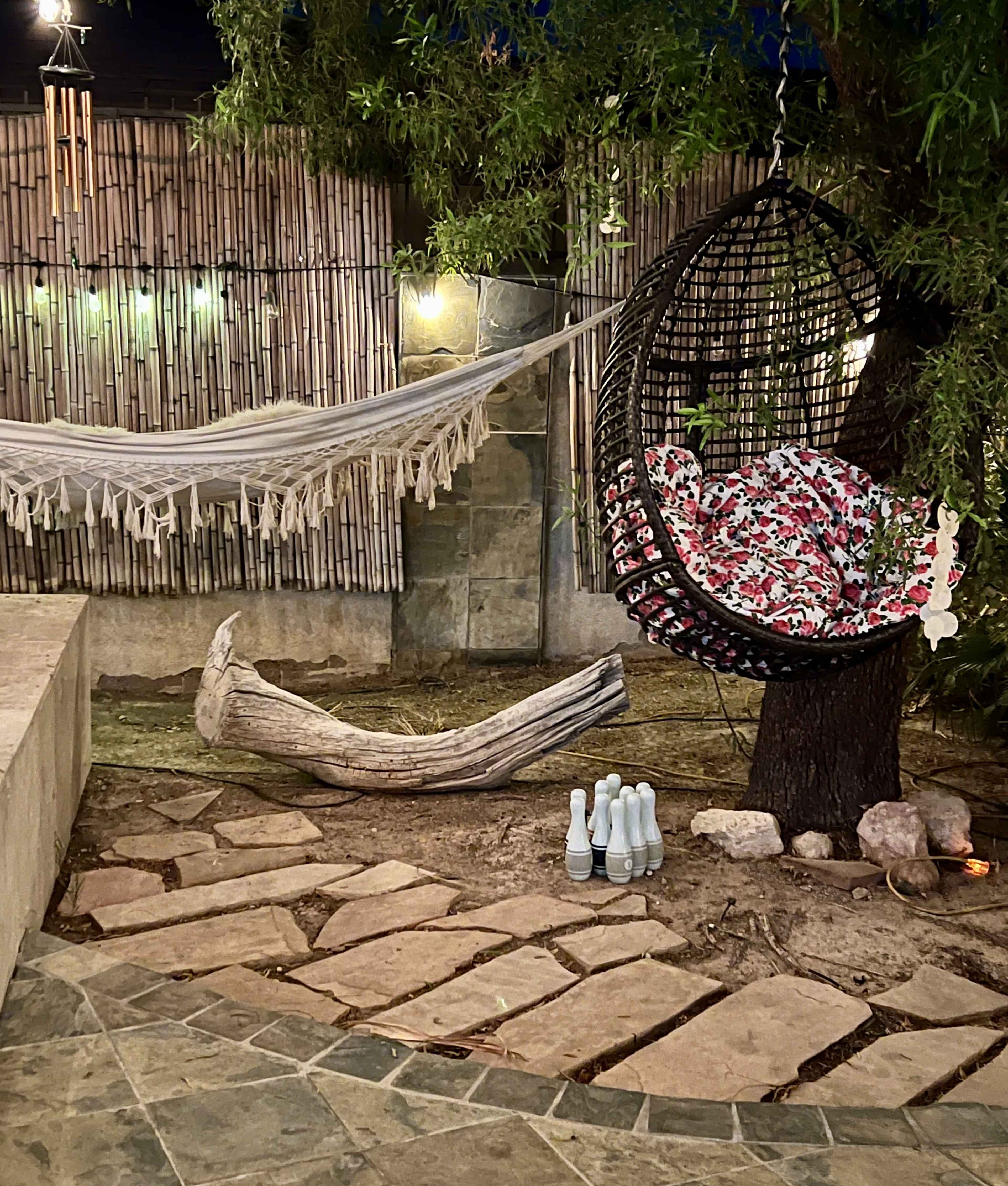 The image shows a cozy outdoor space featuring a hanging chair with floral cushions, a hammock strung between two posts, a rustic log, and a set of light blue bottles arranged on a stone pathway.