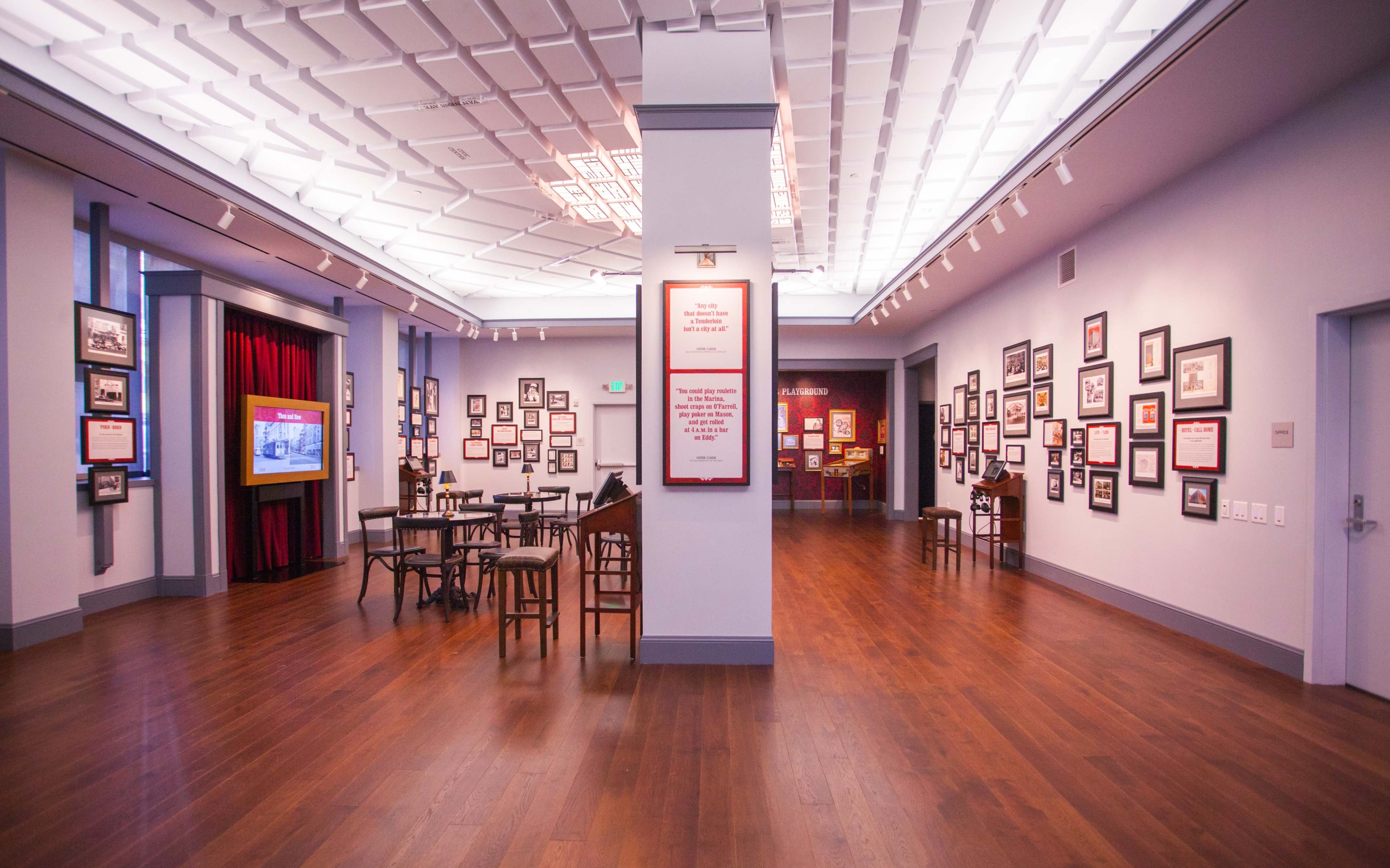 An interior of a museum or gallery with wooden flooring, framed pictures on the walls, and several tables arranged in the space.