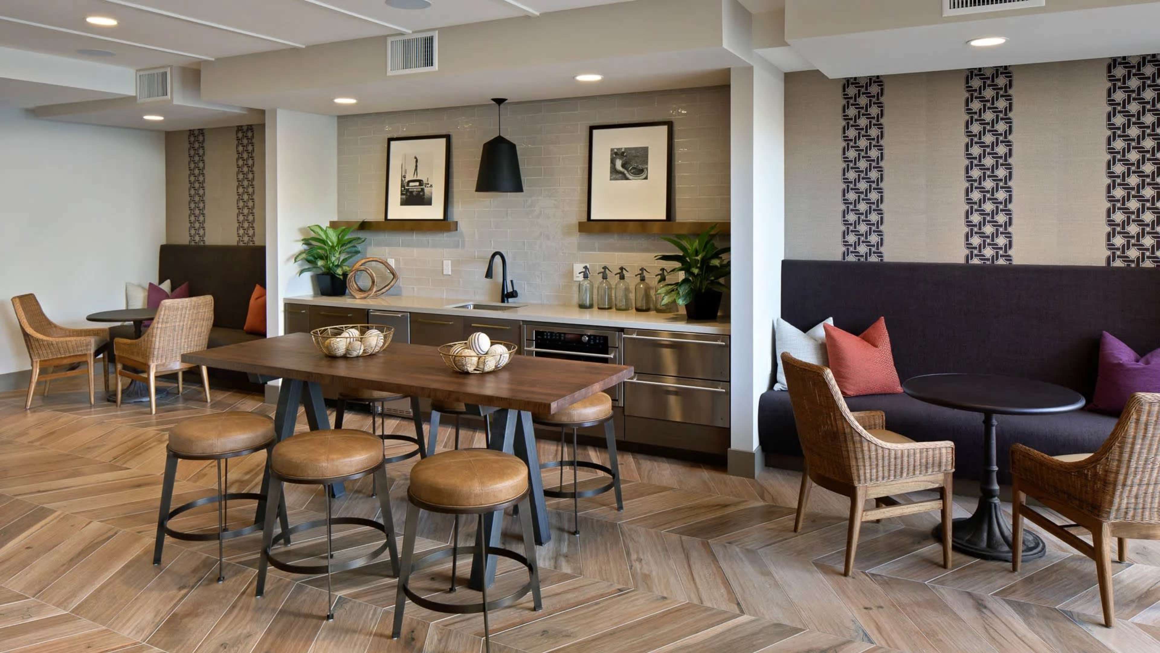 A modern kitchen and dining area features a wooden table with stools, seating arrangements, and decorative plants against a backdrop of stylish wall patterns.