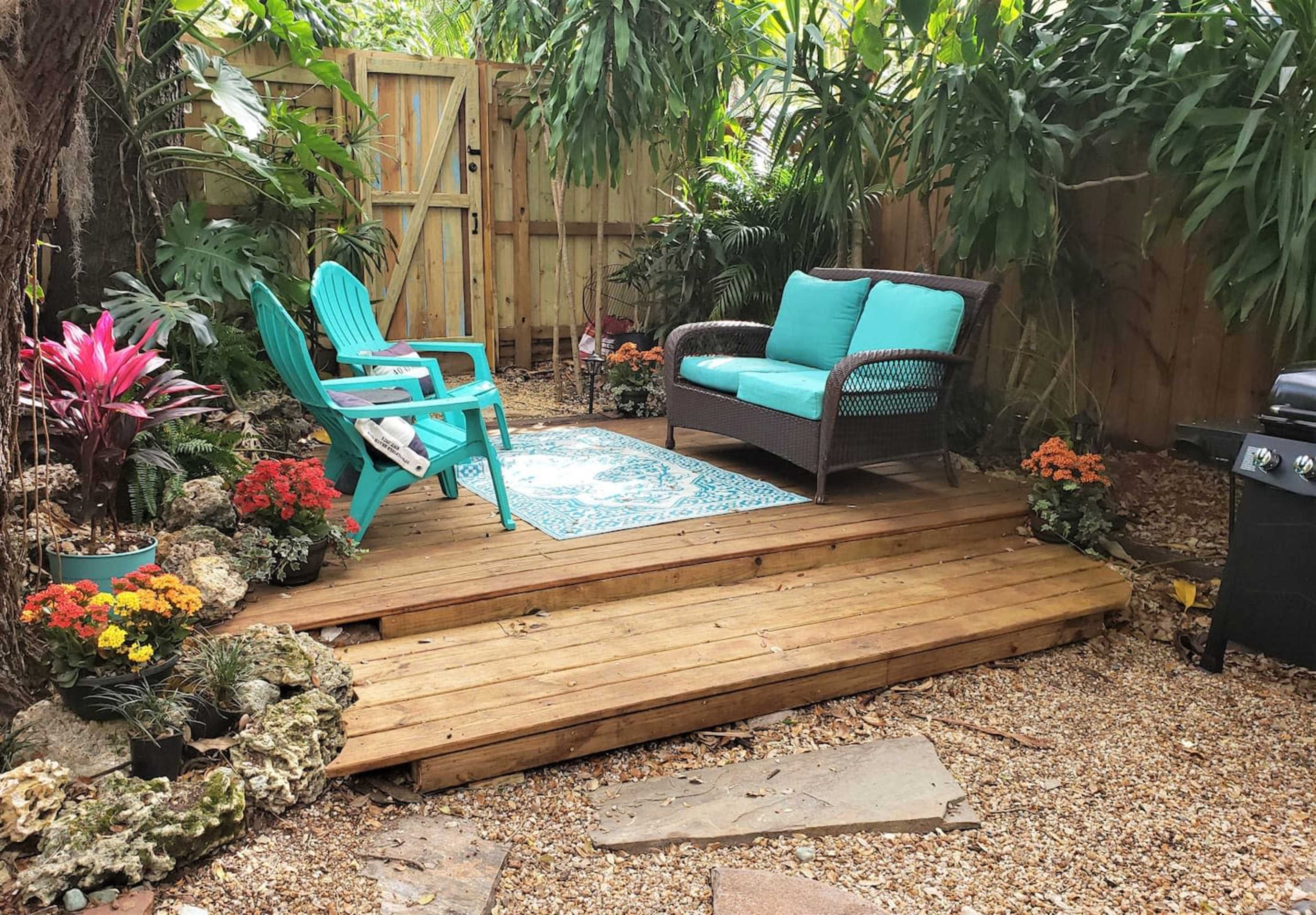 The image shows a wooden deck in a garden area, featuring a turquoise sofa and an Adirondack chair surrounded by potted plants and flowers.