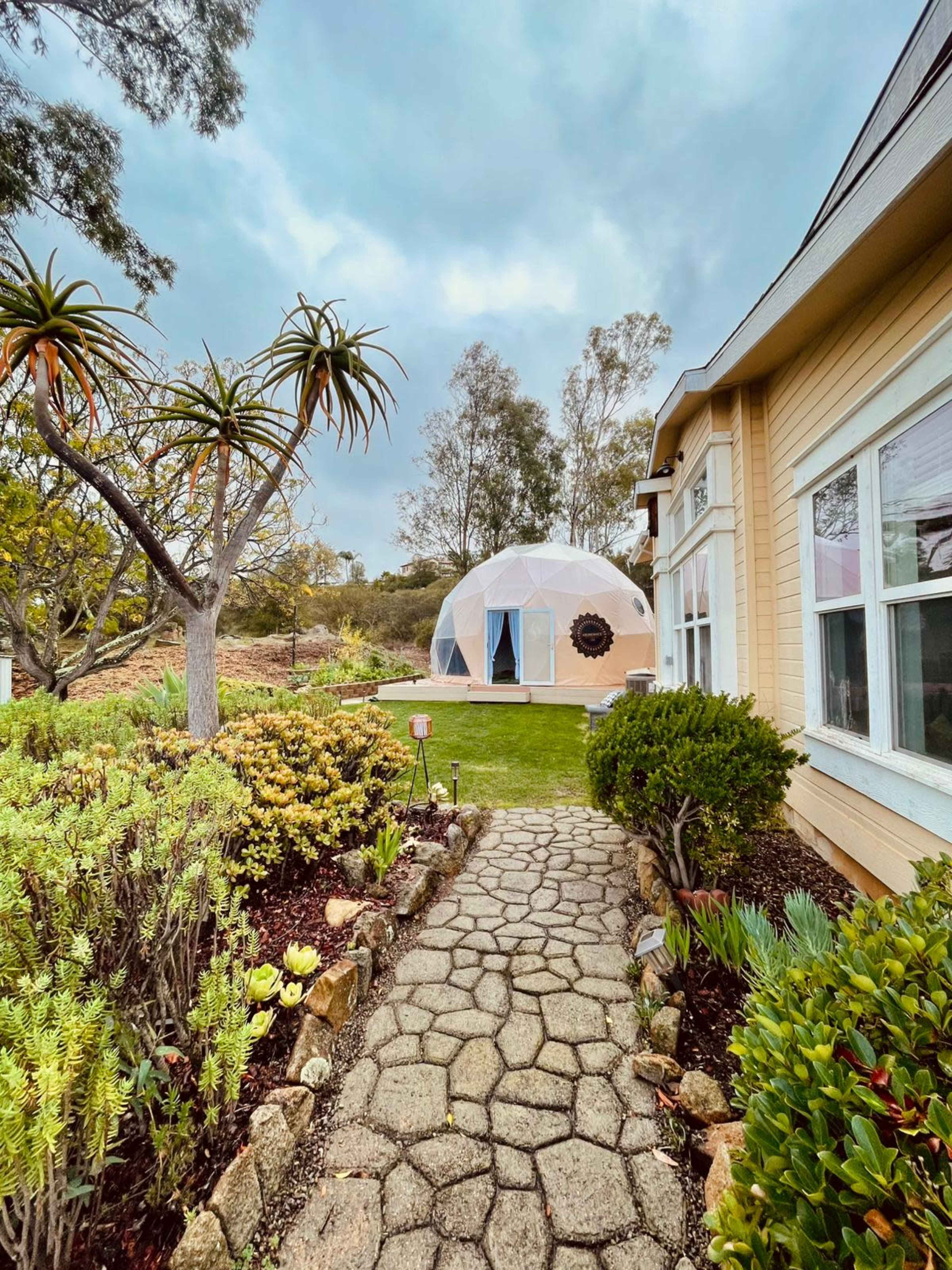 A pathway made of stone leads through a garden to a geodesic dome structure situated near a house.