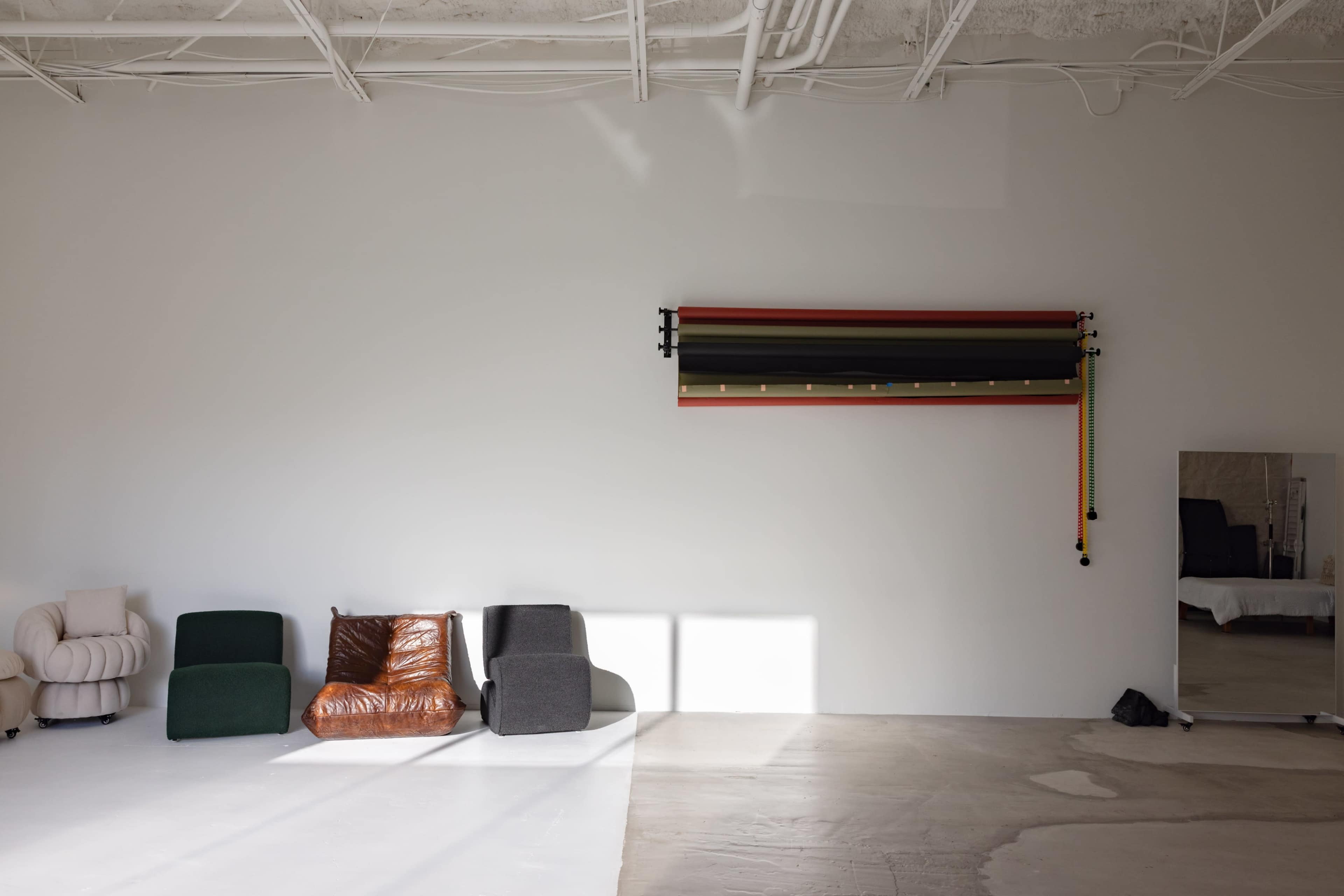 A minimalist interior features three chairs in varying colors and a mounted rack on the wall above, with a mirror reflecting the space.