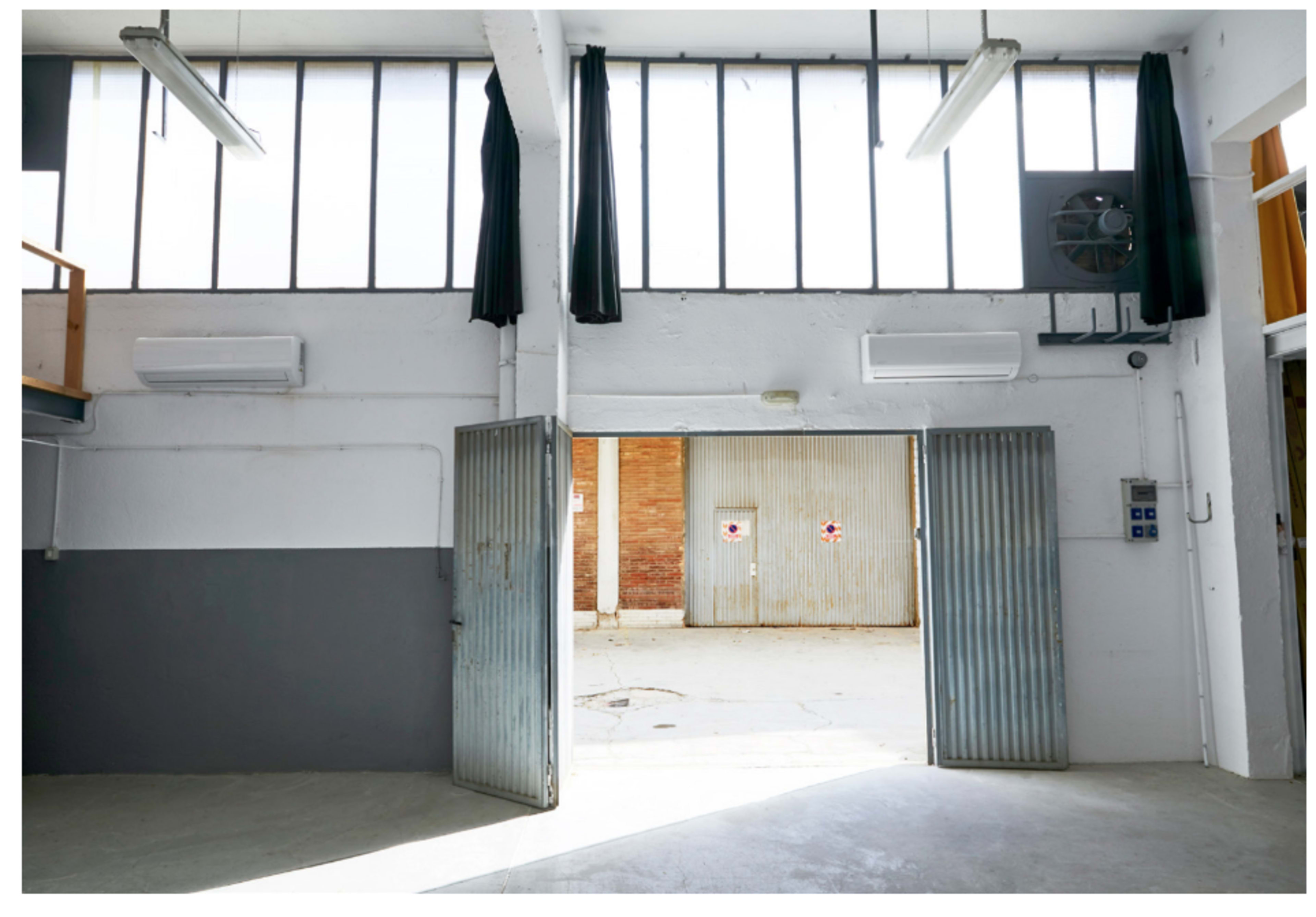 An industrial space with large windows, open double doors, and a view of a brick wall and closed metal shutter beyond.