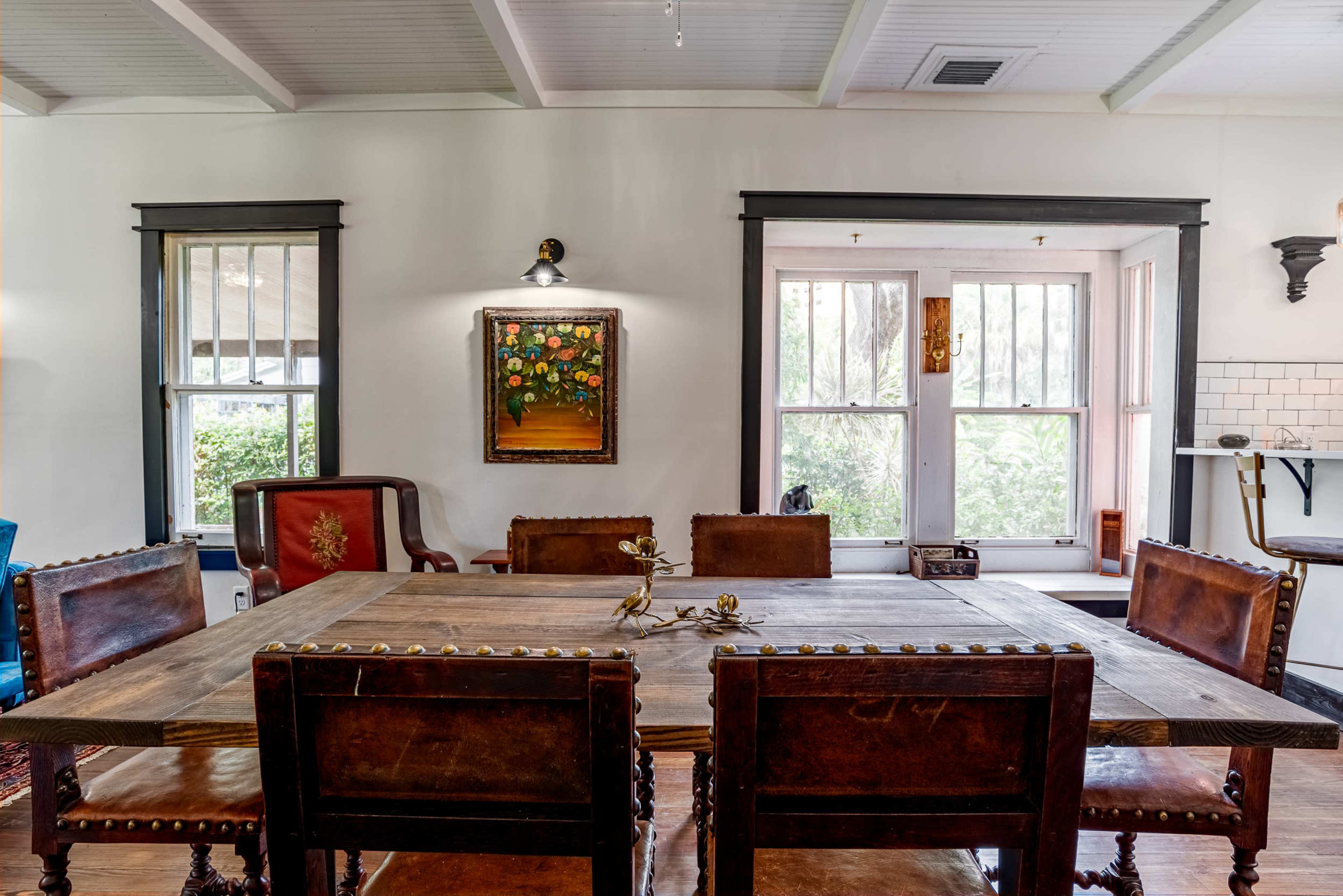 A wooden dining table with six chairs is set in a well-lit room featuring a window and a piece of framed artwork on the wall.