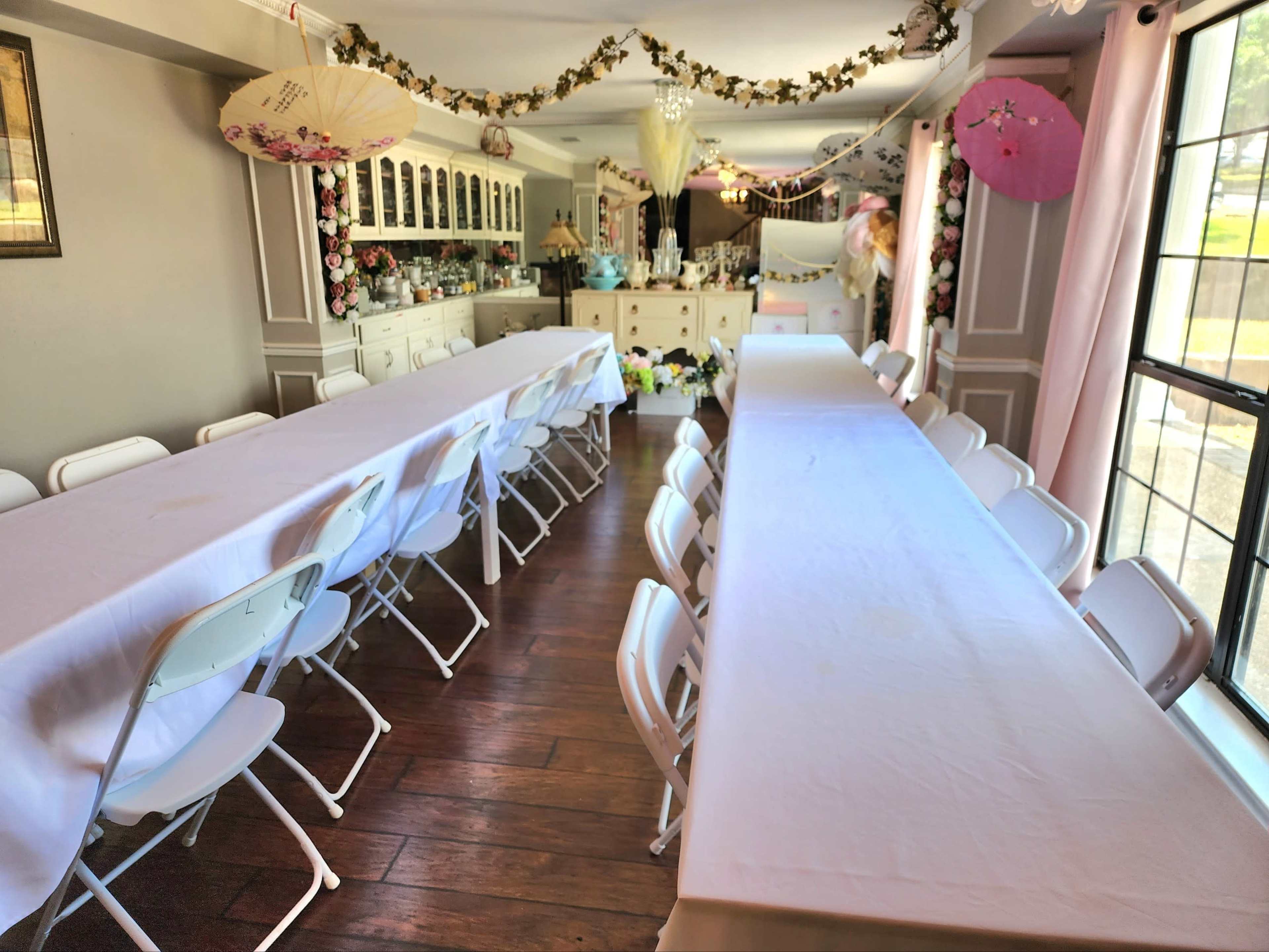 A decorated room with two long tables covered in white tablecloths, arranged with folding chairs along the sides, and floral decorations hanging overhead.