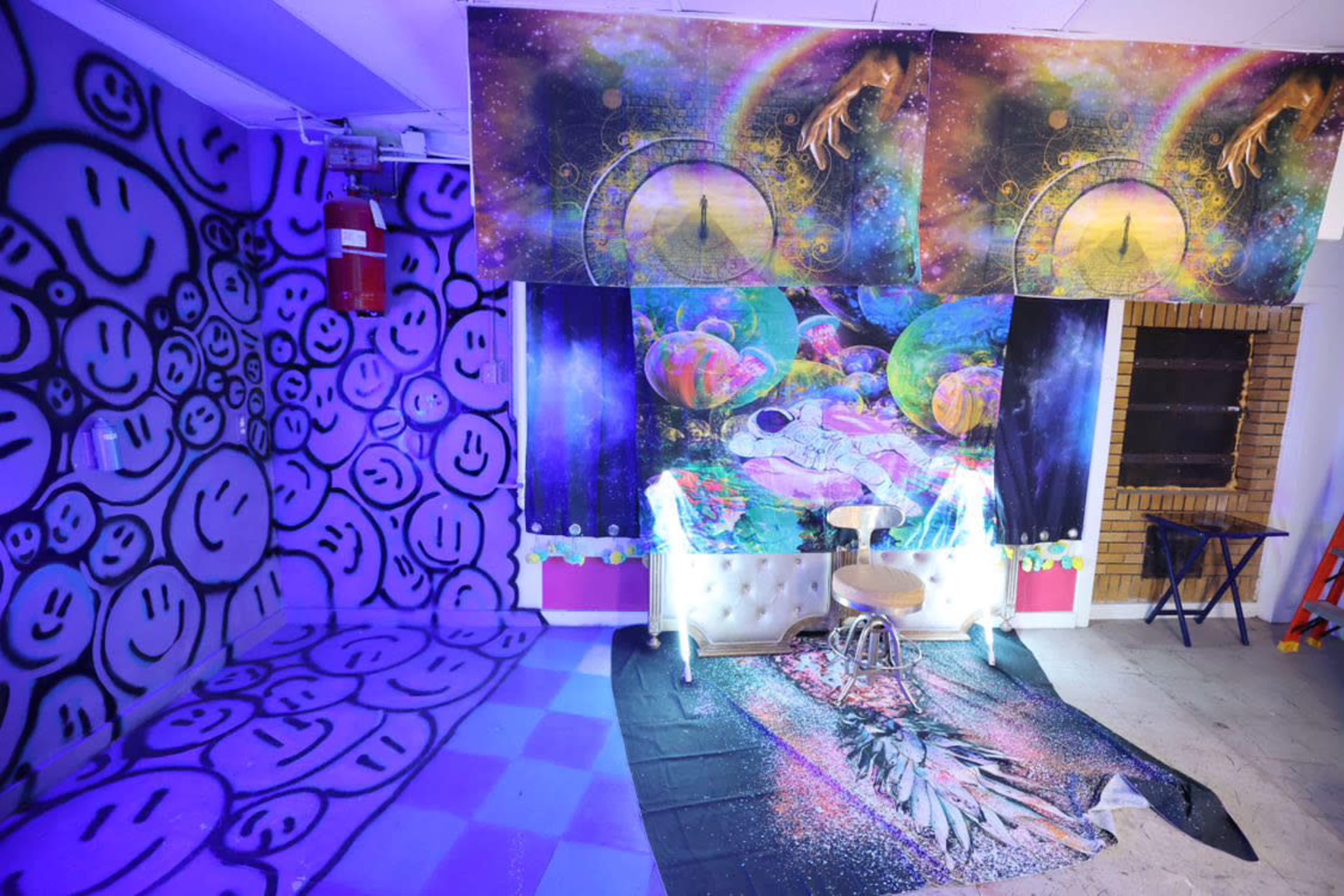 A room decorated with vibrant graffiti of smiley faces on one wall, colorful cosmic-themed art on the ceiling, and a white couch with a stool positioned in front of it.