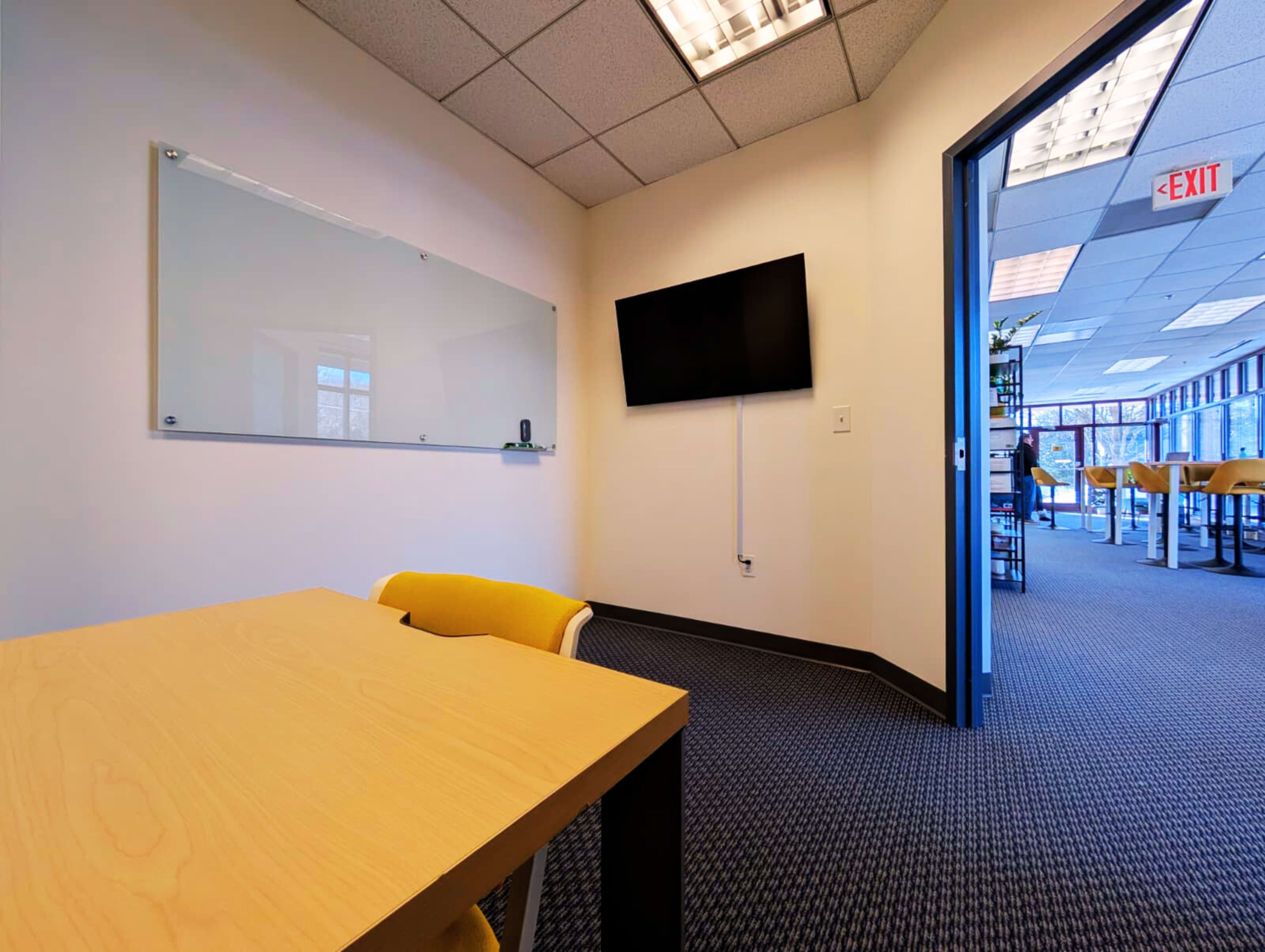 A small meeting room features a light-colored table and yellow chairs, with a whiteboard and a television mounted on the wall, leading to a hallway with additional office space visible.