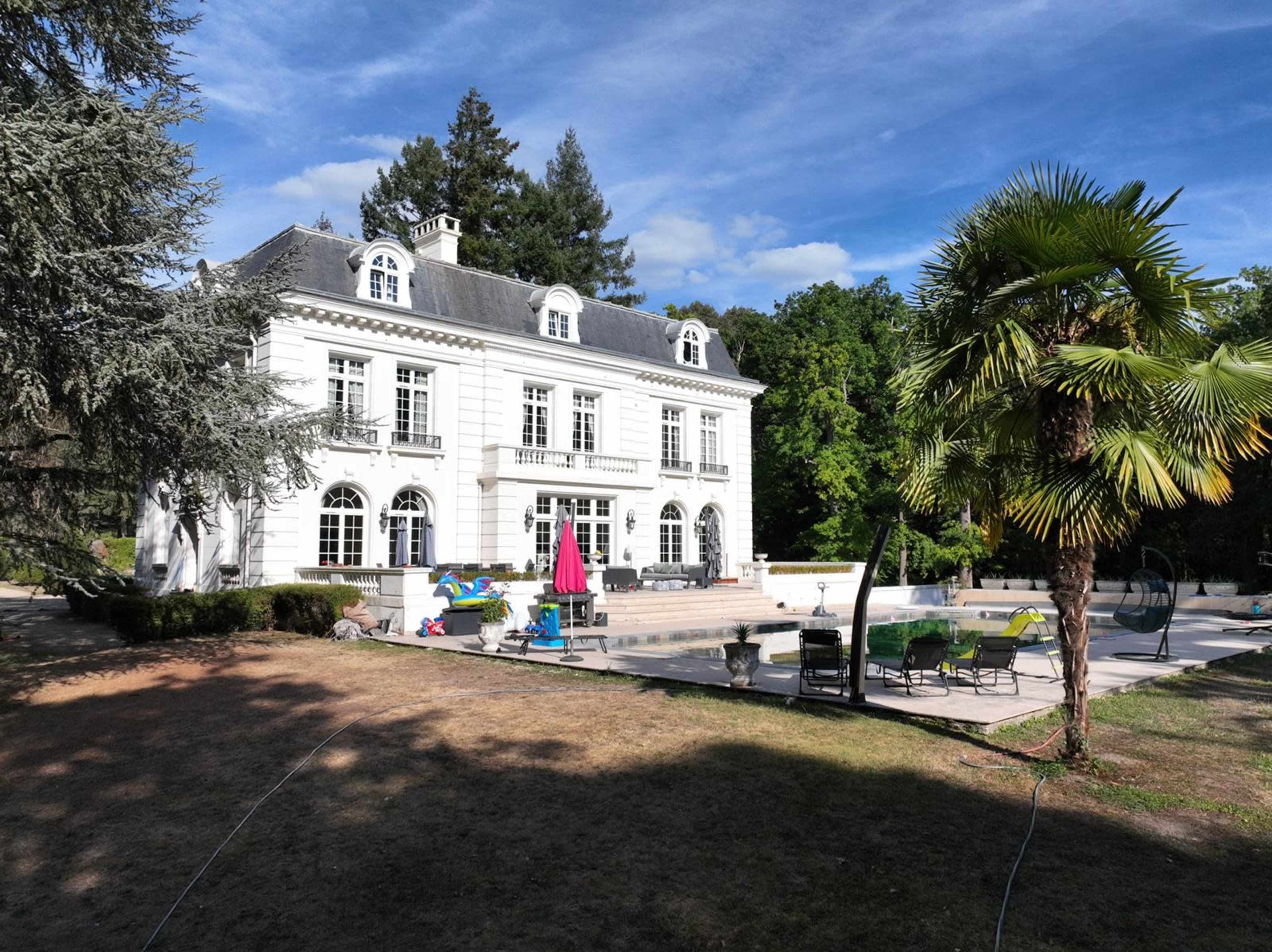 A large, white mansion with multiple windows and a patio area featuring lounge chairs and a swimming pool is surrounded by greenery and palm trees.