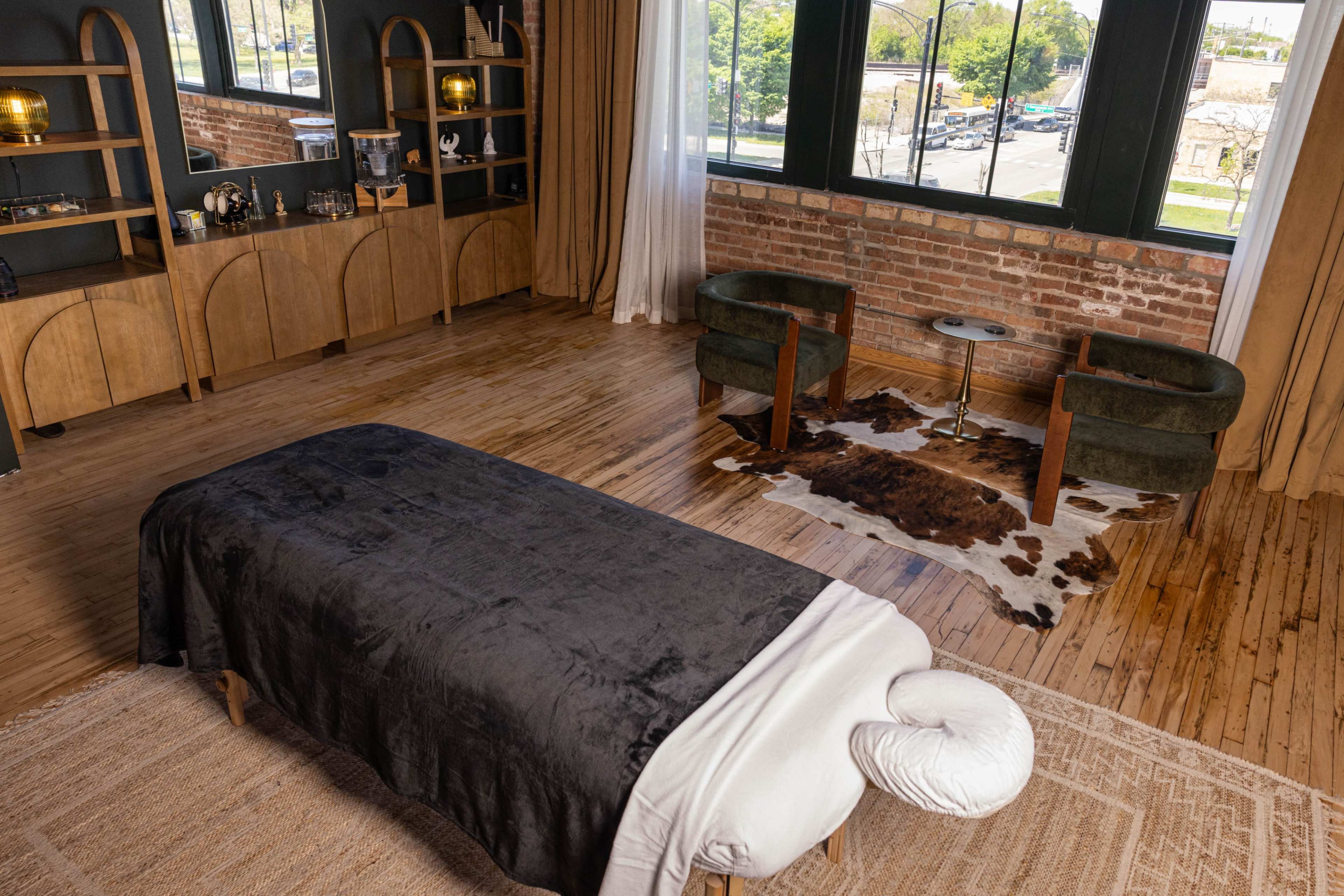 The image shows a massage room featuring a treatment table covered with a black blanket, two green chairs, a decorative cowhide rug, and large windows with a view of the outside.