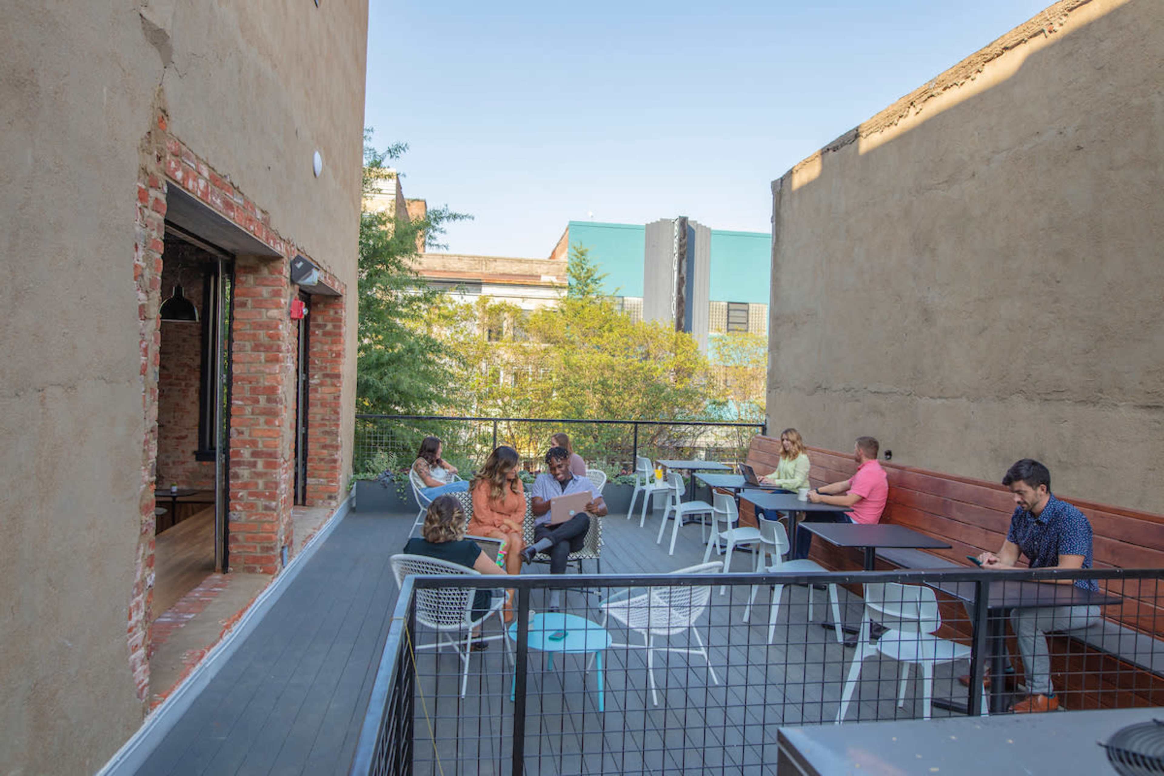 A rooftop café features several individuals seated at tables, with a view of surrounding buildings and greenery.