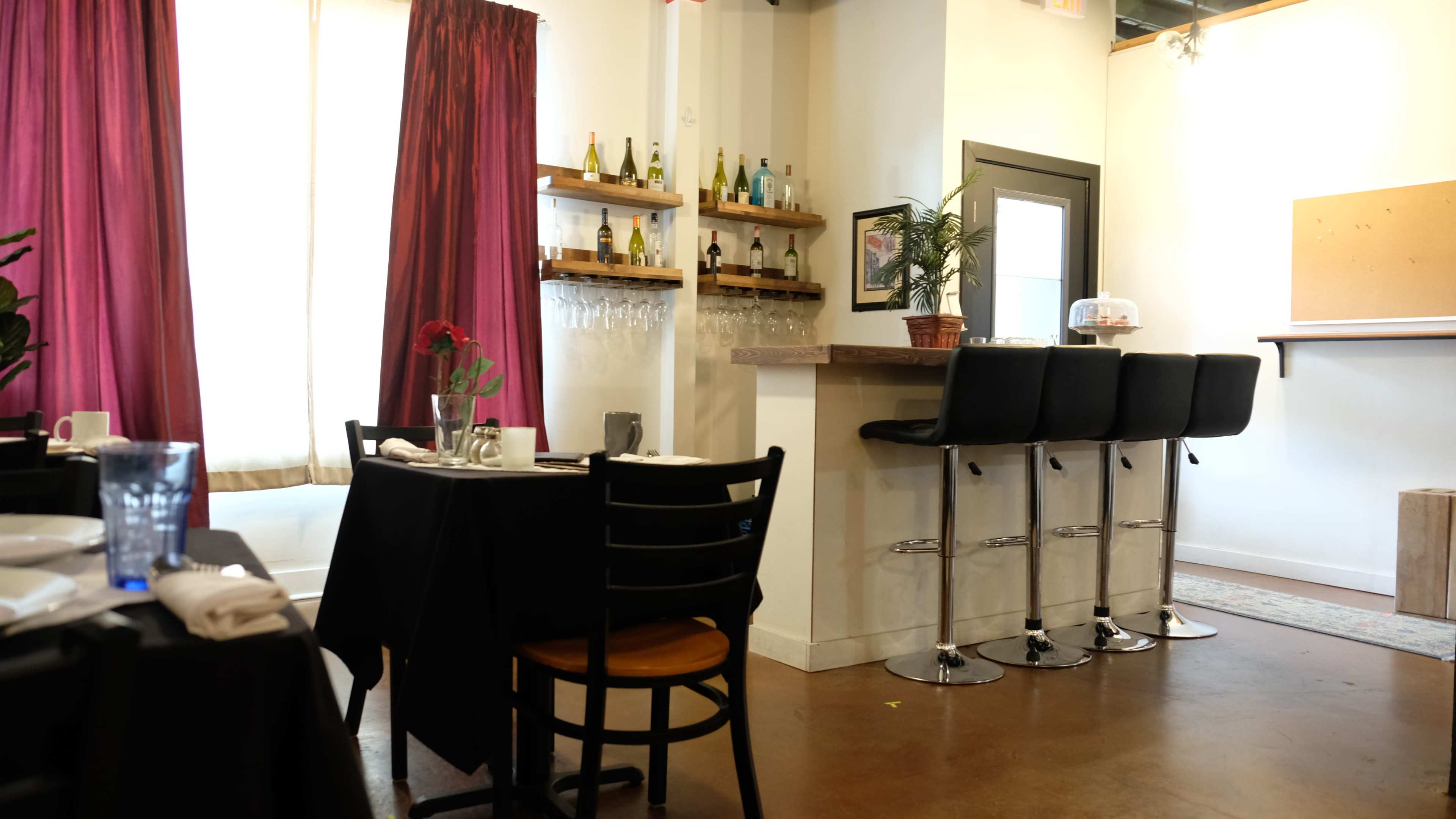 A restaurant interior featuring black tables, bar stools at a counter, and curtains in shades of red.