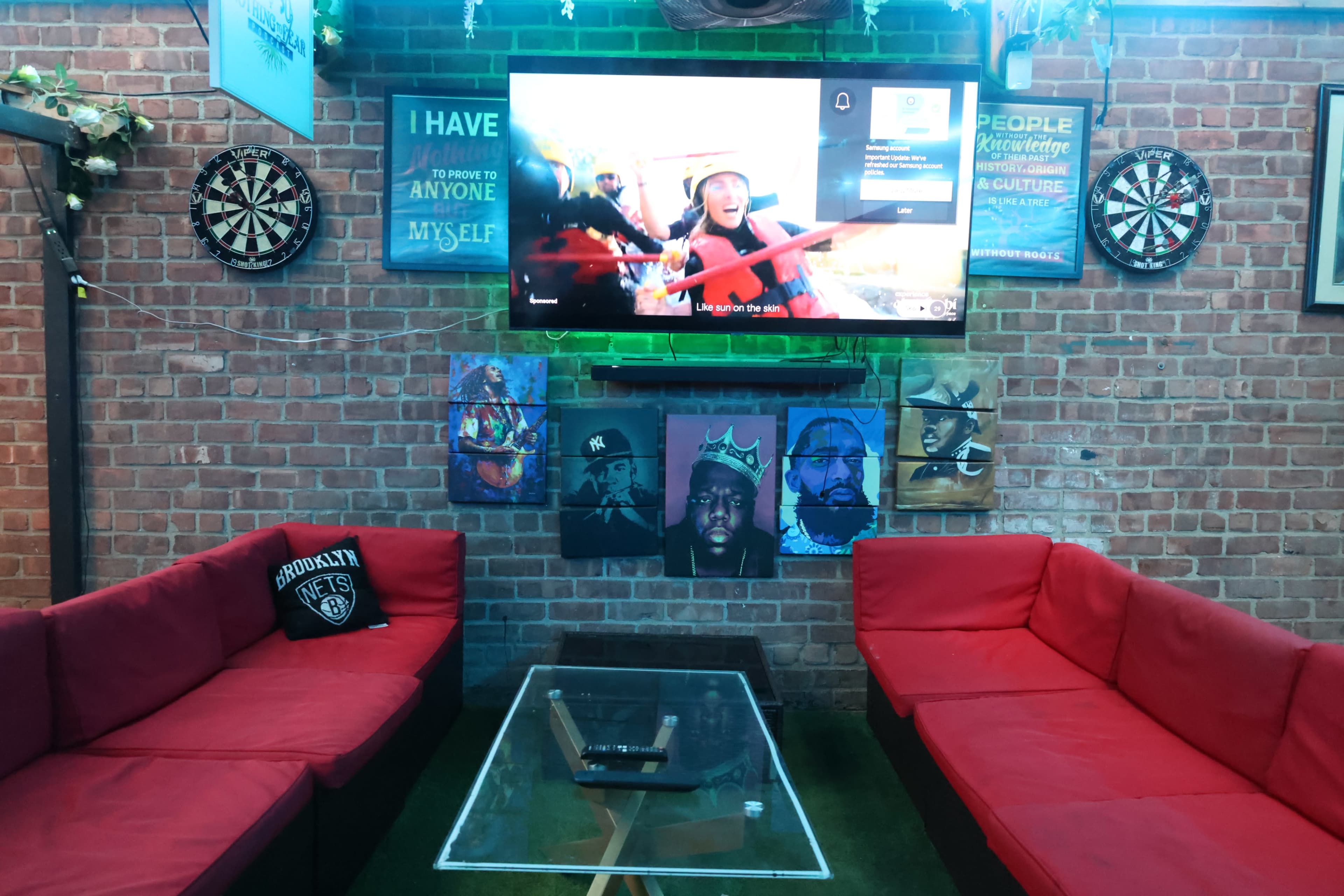 The image shows a cozy lounge area with red couches, a glass coffee table, and a wall adorned with artwork and a television displaying a program.