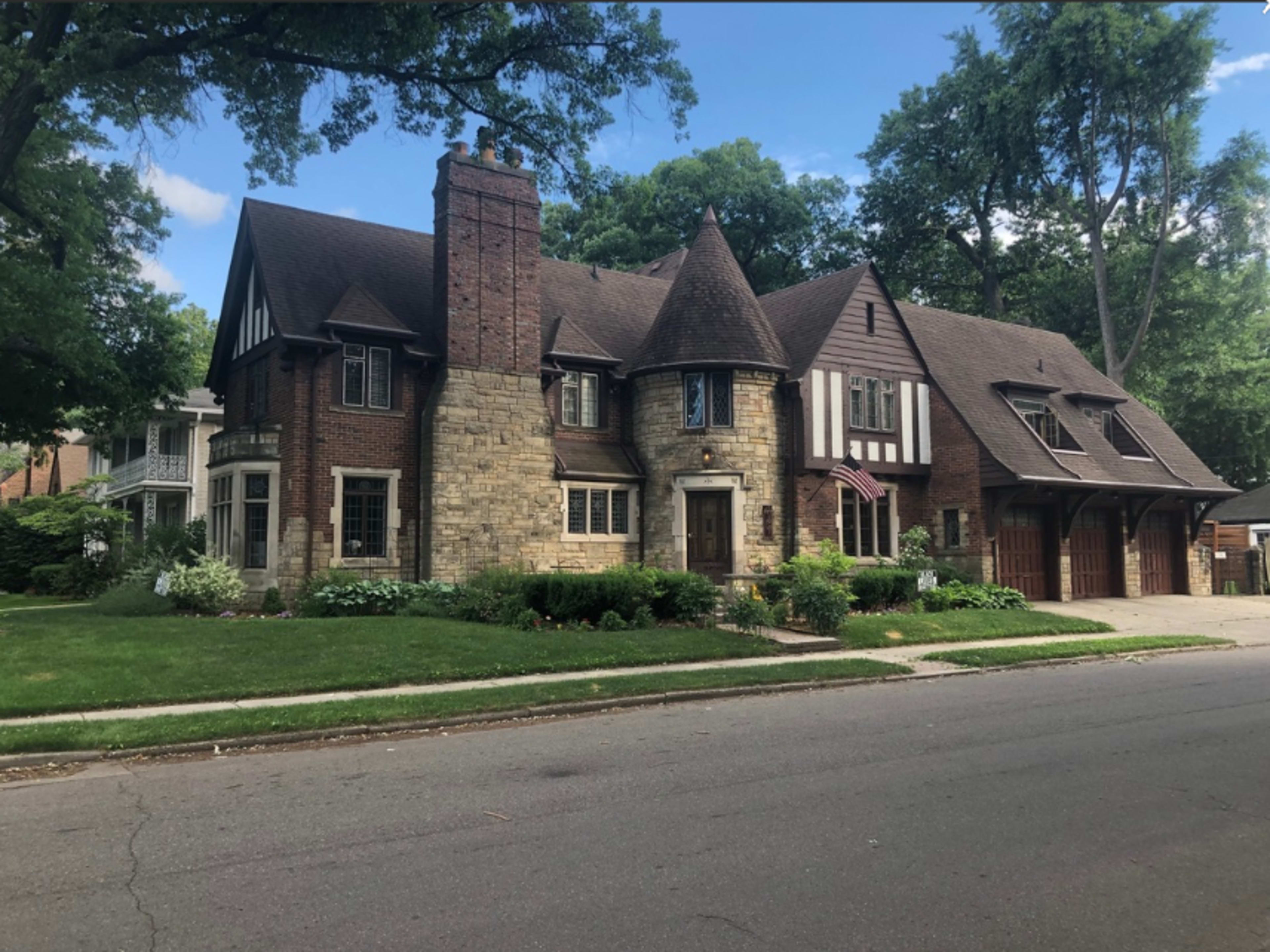 A large, two-story, Tudor-style house with a combination of brick and stone exteriors, featuring a prominent chimney and multiple gabled roofs.