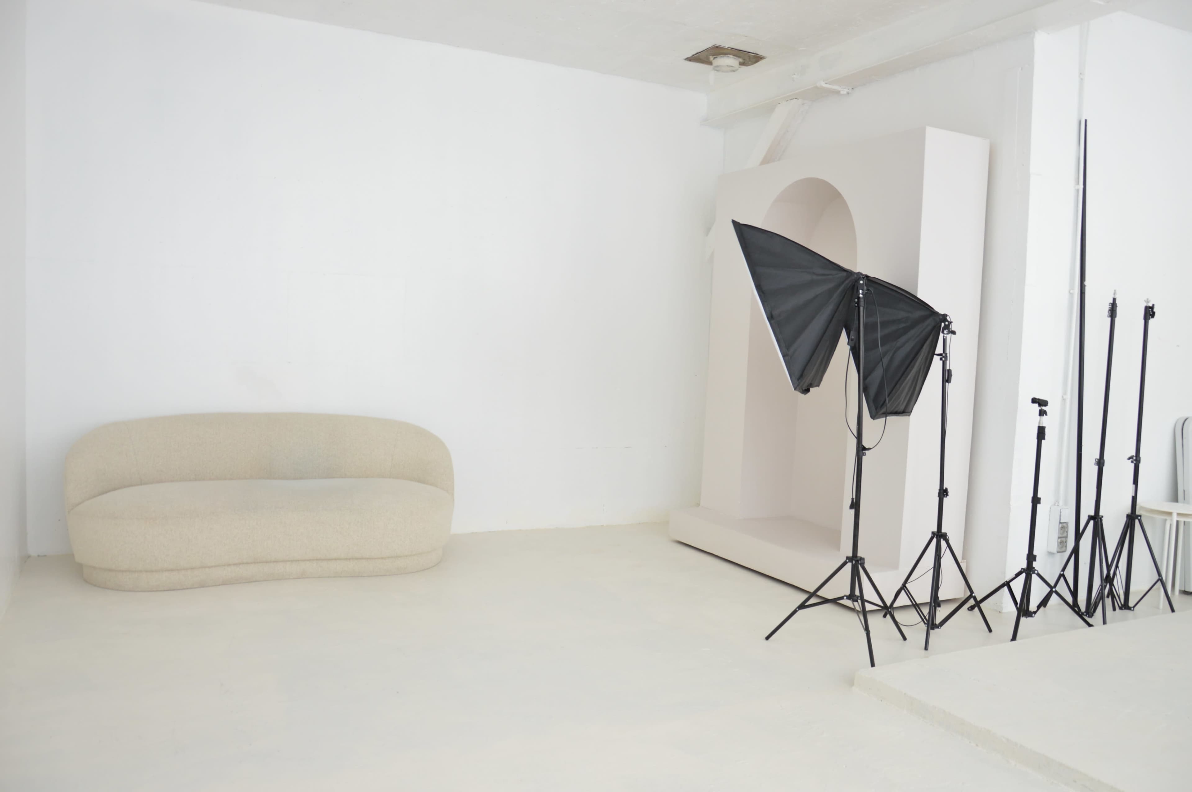 The image shows a minimalist interior with a curved beige sofa, a sculptural white alcove, and a photography setup including a large softbox and light stands.