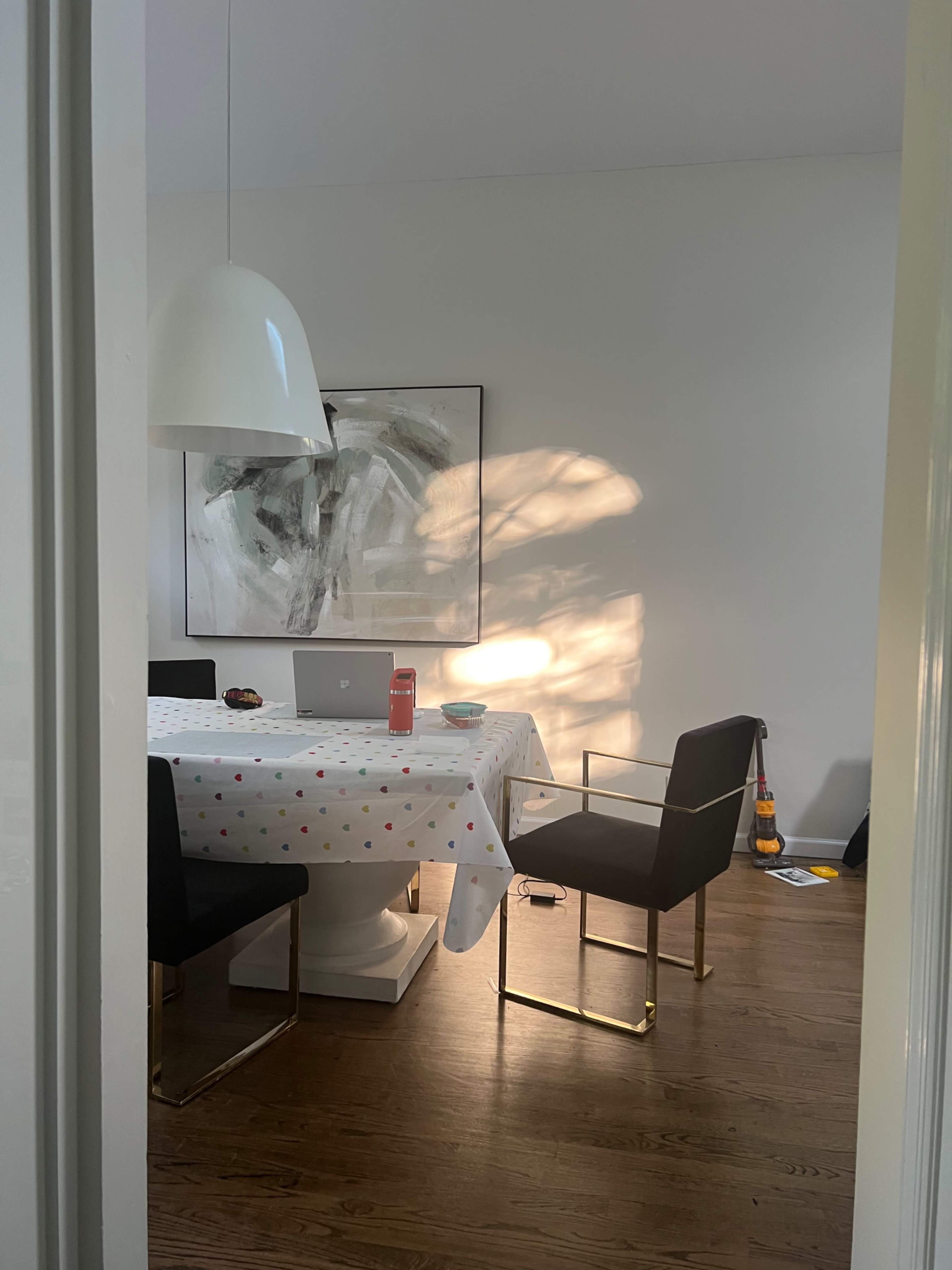 A dining area features a white table with a polka dot tablecloth, surrounded by modern black chairs, and a large pendant light overhead, with sunlight casting shadows on the wall.
